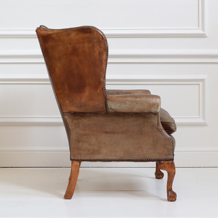 A vintage wingback armchair with a Queen Anne influence given by the cabriole front legs and carved wooden feet. The distressed brown leather upholstery highlights the natural wear and a lovely aged character. The deep button tufting on the inside