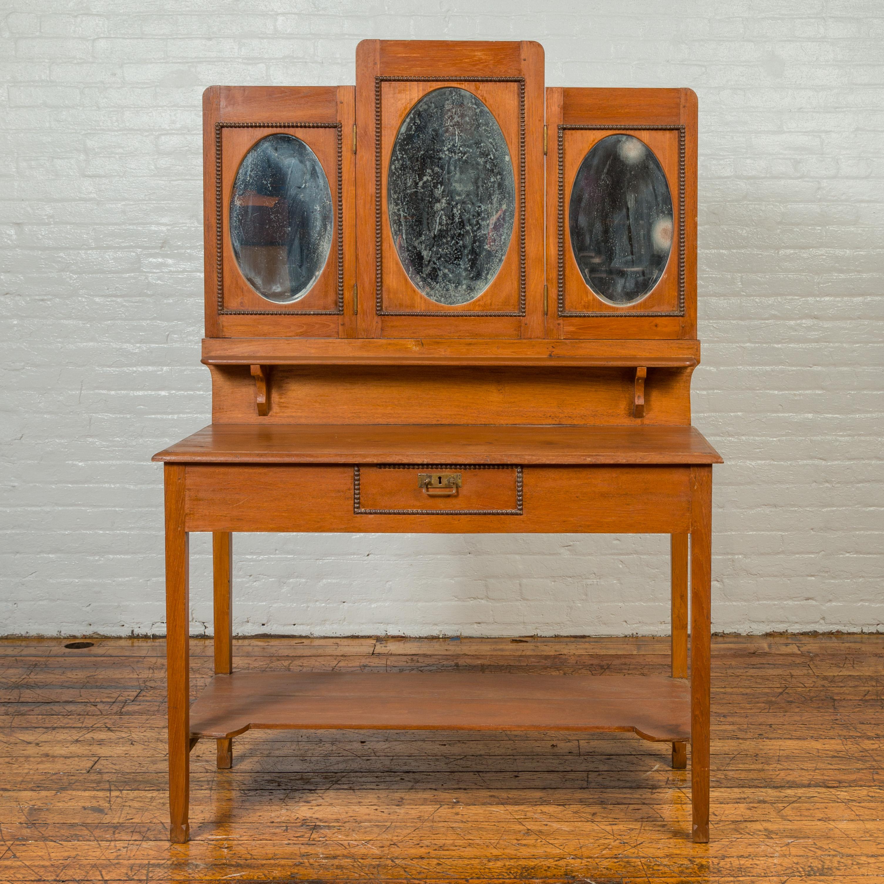 A vintage Indonesian woman's vanity from the mid-20th century, with oval glass panels, single drawer and lower shelf. Crafted during the midcentury period, this wooden vanity features a central door panel flanked with folding ones, smaller in size.