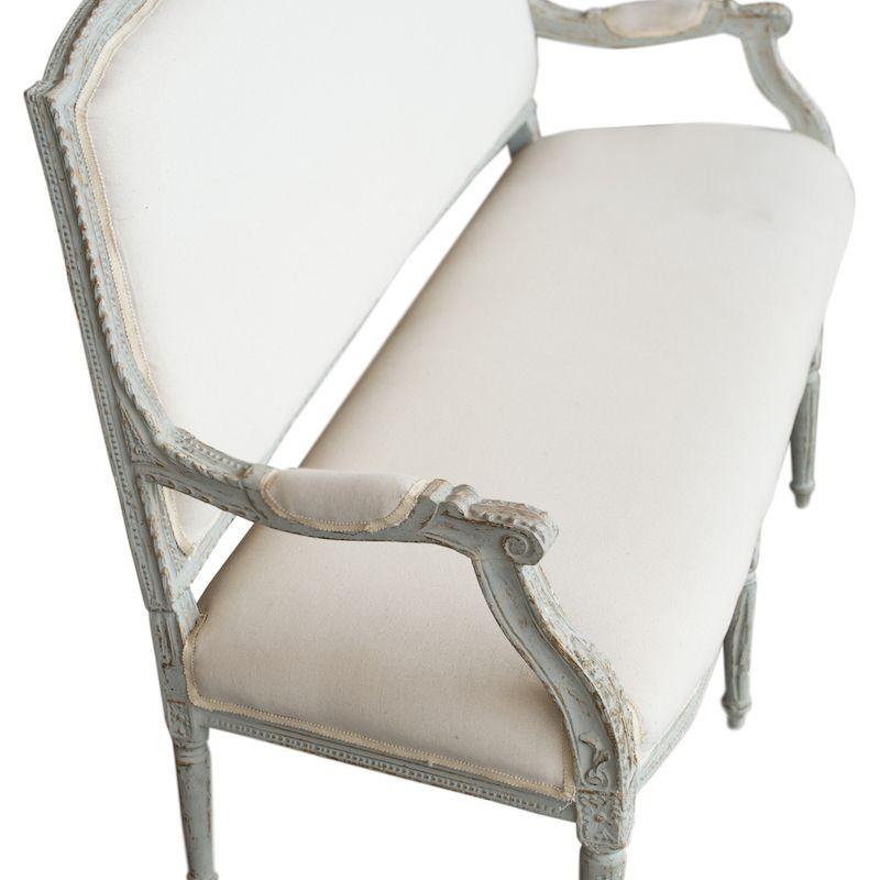 Un canapé classique en bois vintage avec des détails ornés et des coussins en lin crème. Le cadre en bois, simple mais détaillé, est peint d'une couleur bleu-gris clair, qui s'est écaillée et usée avec le temps. Les détails sculptés sont simples
