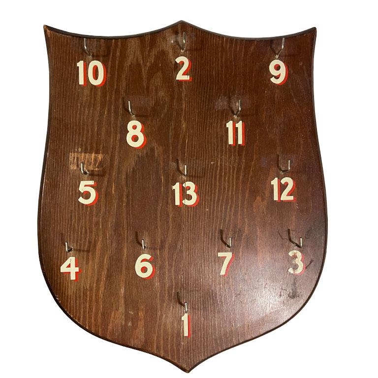 Un tavolo da gioco vintage in legno per il lancio degli anelli o un portachiavi per hotel. Questo pezzo si rifà ad Abercrombie & Fitch ed è realizzato in legno. Ha la forma di uno scudo e i bordi smussati. Su tutta la superficie sono applicati