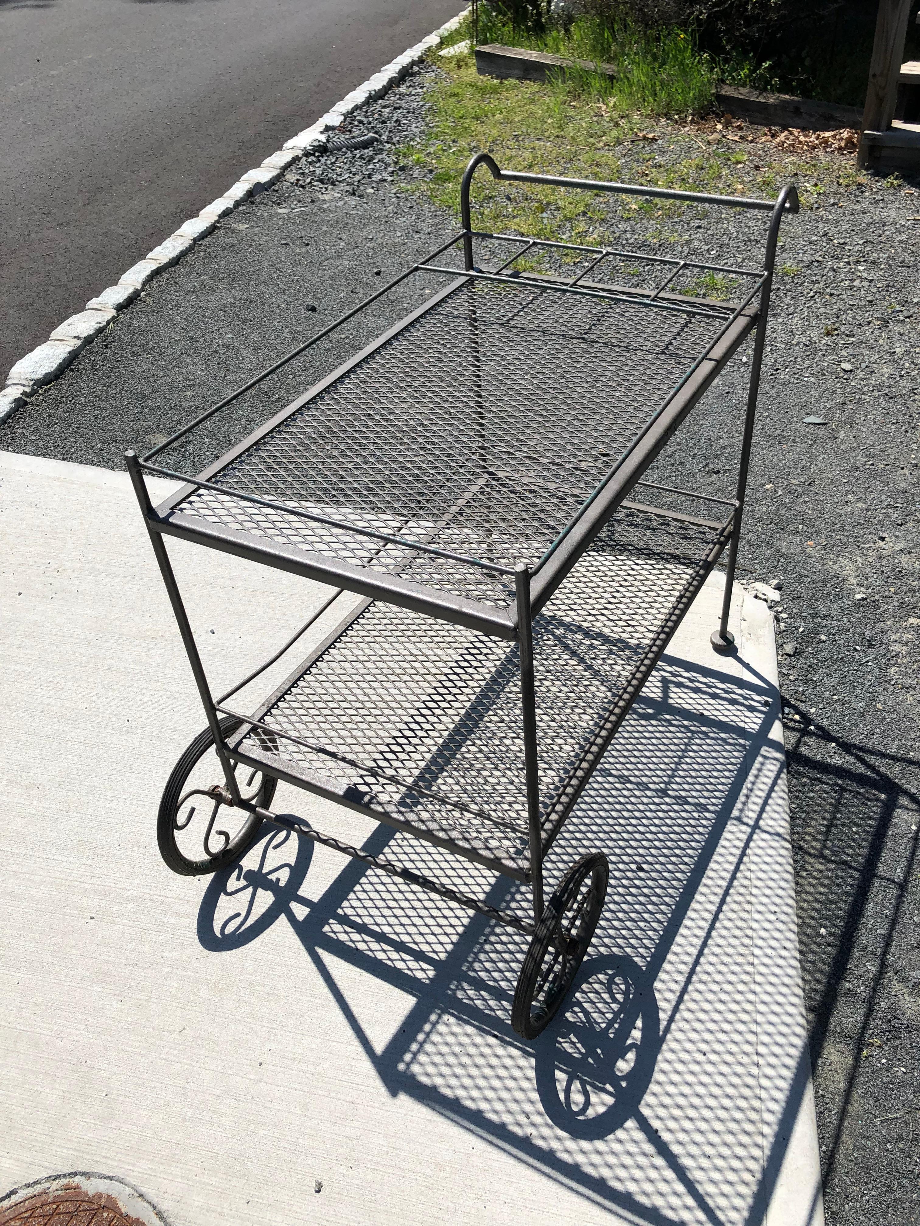 Classic vintage Woodard outdoor iron bar cart painted a gun metal color with some visible green undercoat. The cart has rubber tires, two tiers, and galleries around each surface. The top tier has space for bottles.
30.5 h to handle
27 h to