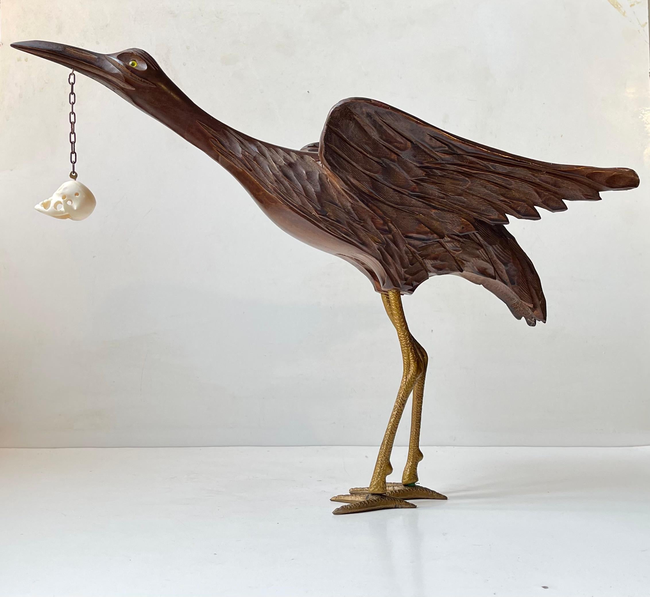 Majestic semi-peculiar and possibly up-cycled/repurposed sculpture. It depicts a detailed crane in carved dark brown wood with glass eyes holding a suspended skull in white bone from its bea. It legs are made from molded bakelite in a golden