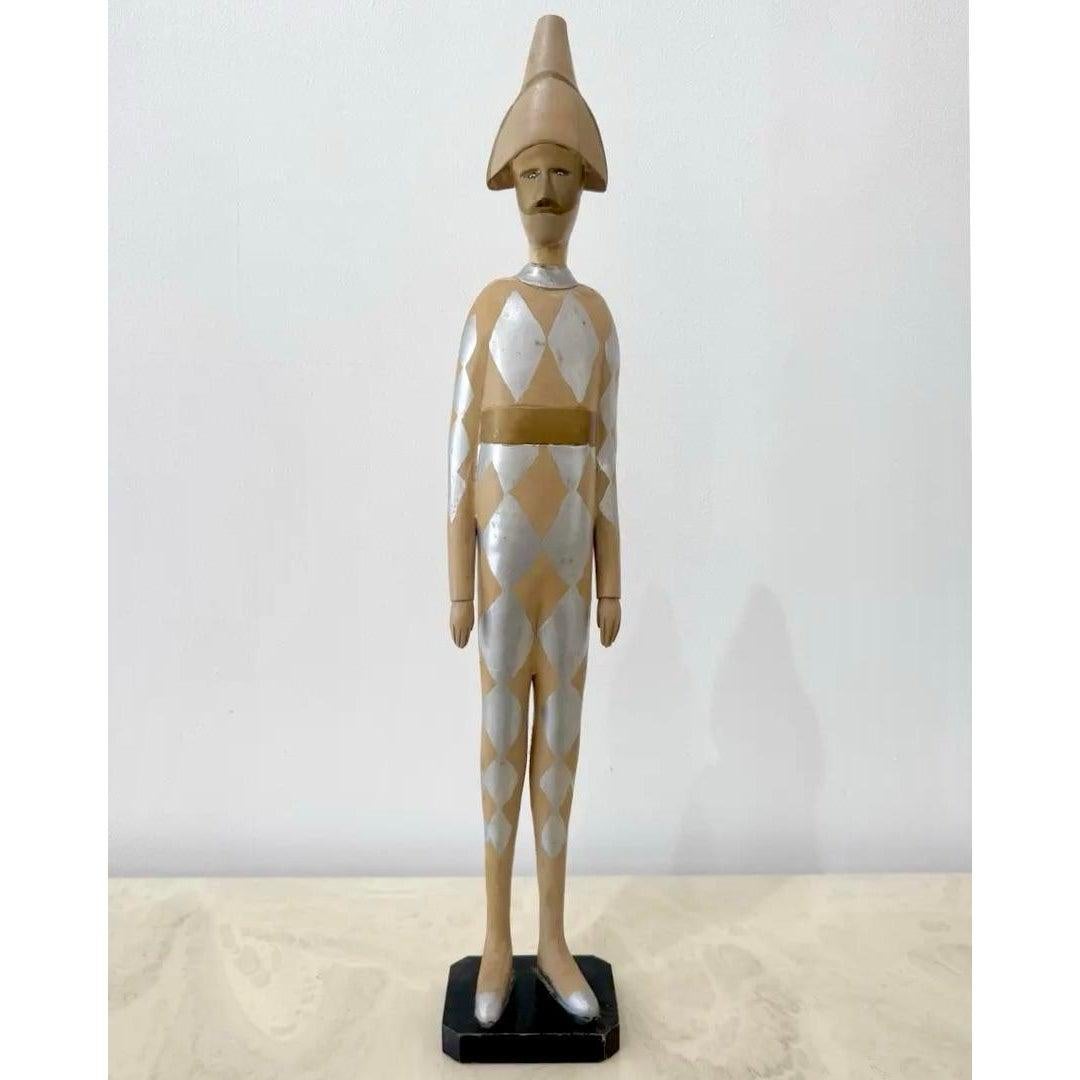 Scultura vintage in legno di un arlecchino stoico, vestito con un body argentato e ocra a rombi e con un cappello a punta abbinato. La forma spigolosa e l'espressione muta le conferiscono un'energia ossessionante, quasi cerimoniale: una via di mezzo