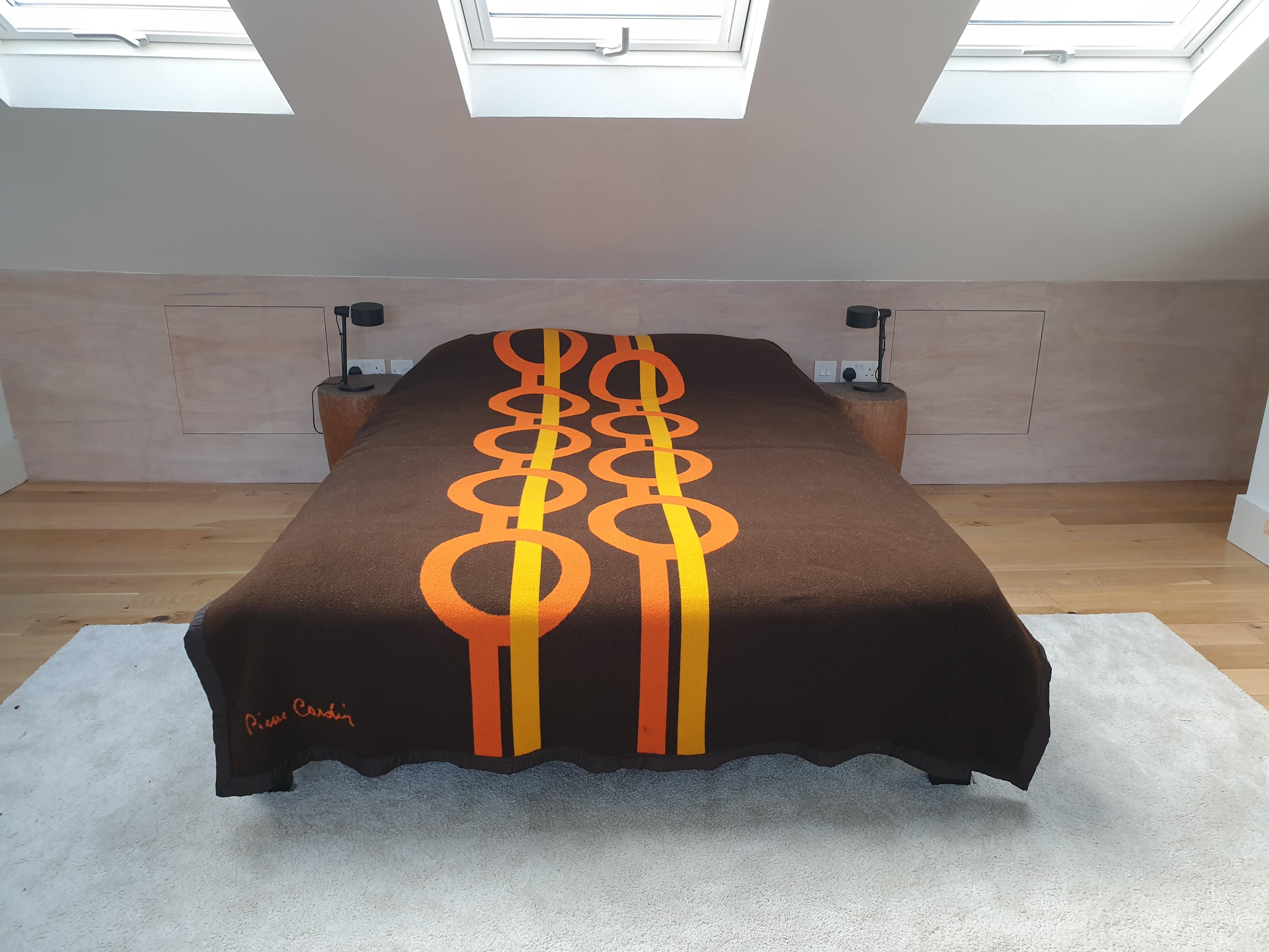 Beautiful woollen bedcover by Pierre Cardin. This cover has the typical intelocking ring design of his work. The main background colour is brown with a design in orange, yellow and black. The edges are bound with a silky edging. The reverse of the