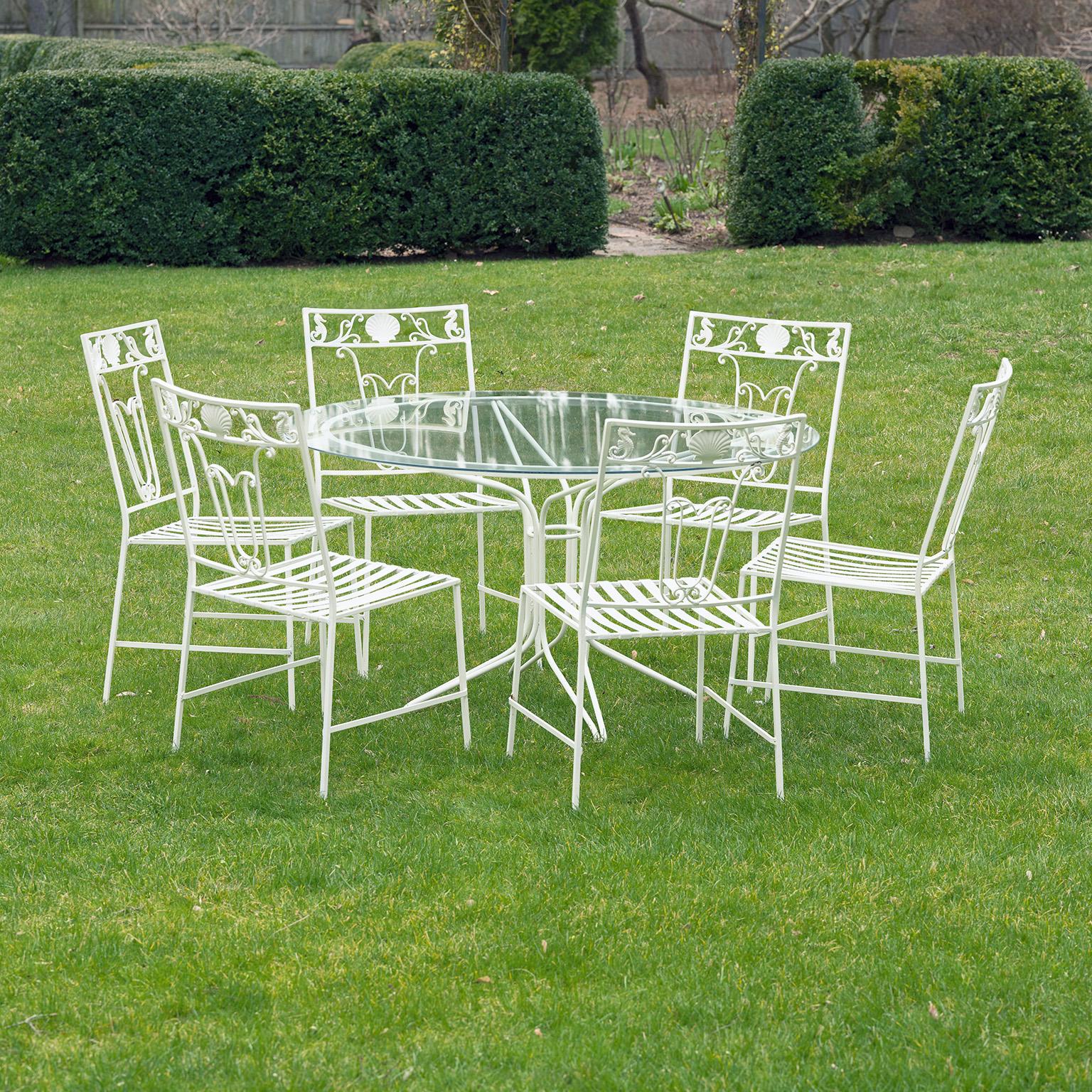 A dining set comprised of a simple wrought-iron table with circular glass top together with six wrought-iron side chairs, the back of each chair with tulip-form scrollwork and a frieze of a single central scallop shell flanked by seahorses. Chair 34