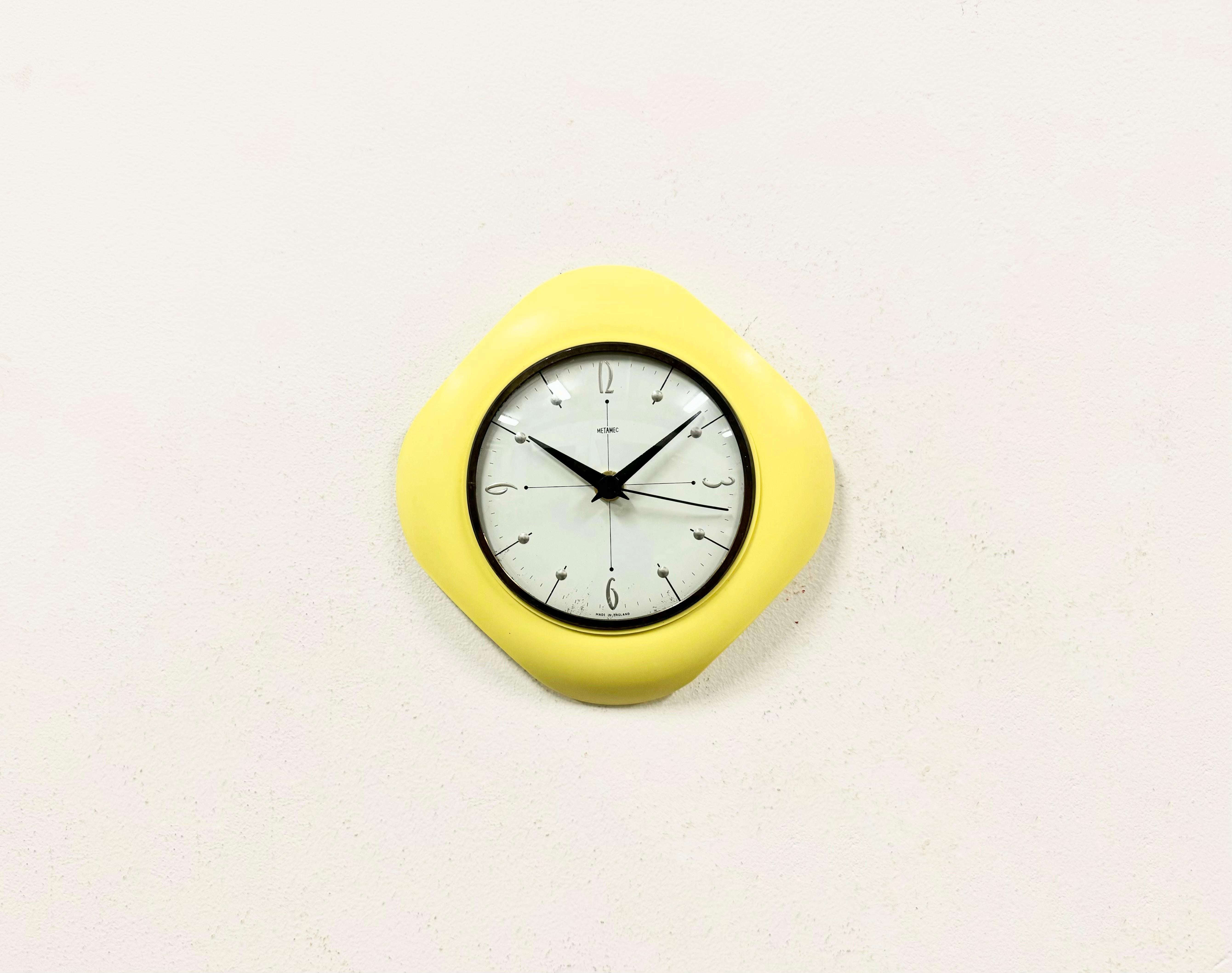 Metamec wall clock made in United Kingdom during the 1970s. It features a yellow bakelite body and a curved clear cover with brass ring. The piece has been converted into a battery-powered clockwork and requires only one AA-battery. The weight of