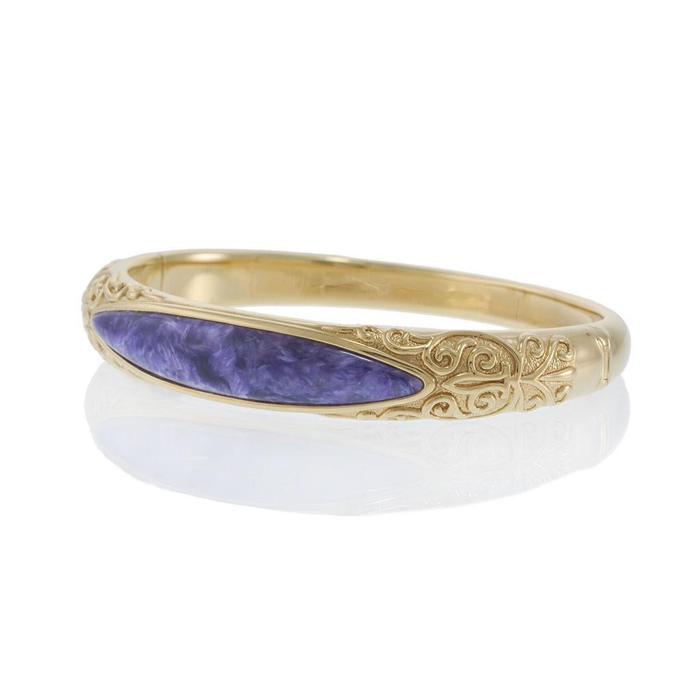 A striking vintage bangle in 18KT yellow gold featuring intricate scrollwork along the band. At its center rests a polished charoite stone, known for its swirling violet hues and unique patterns. The detailed goldwork paired with the rare gemstone