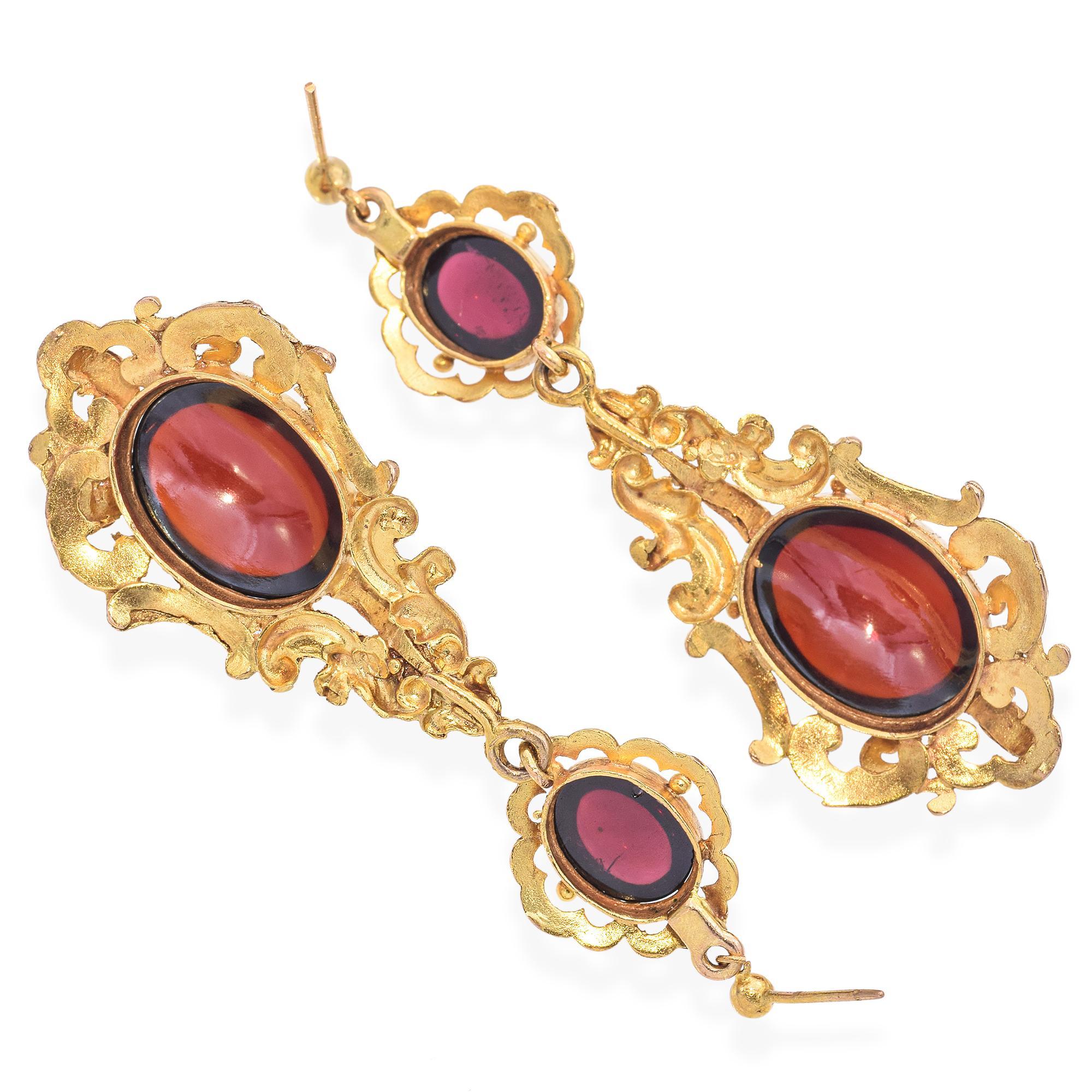 Une paire de boucles d'oreilles exquise avec des pierres cabochons en grenat vibrant. Ces pièces remarquables sont façonnées dans un or riche, formant un cadre complexe et orné d'élégantes volutes. Chaque boucle d'oreille est ornée d'un grenat ovale