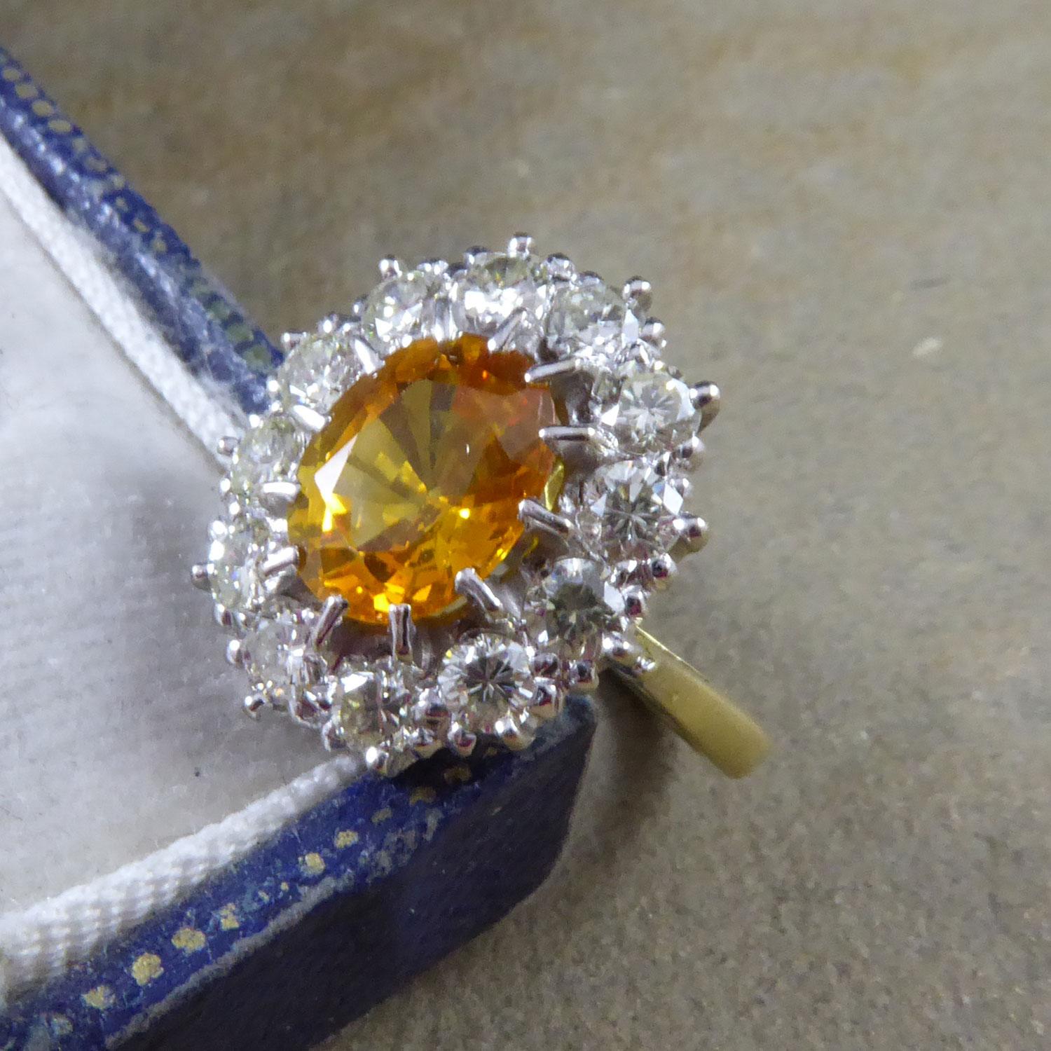 A yellow and white stone set cluster ring centring on a claw set, oval-shaped, mix-cut sapphire, measuring approx. 8.30mm x 6.65mm x 3.88mm deep.  The sapphire sits within a surround of 12 claw-set brilliant cut diamonds, measuring approx.