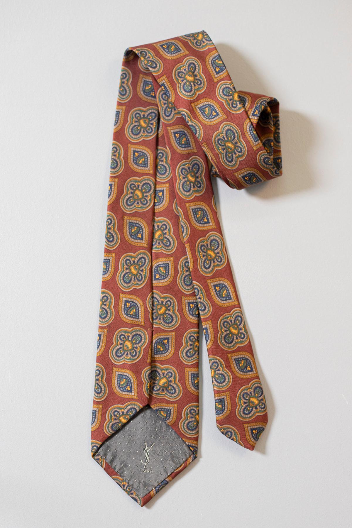 Decorated with blue and yellow paisley motifs on a dark red background, this vintage tie has been designed by Yves Saint Laurent. Made in all silk, this tie is a classic and goes perfectly with every monochromatic outfit. 