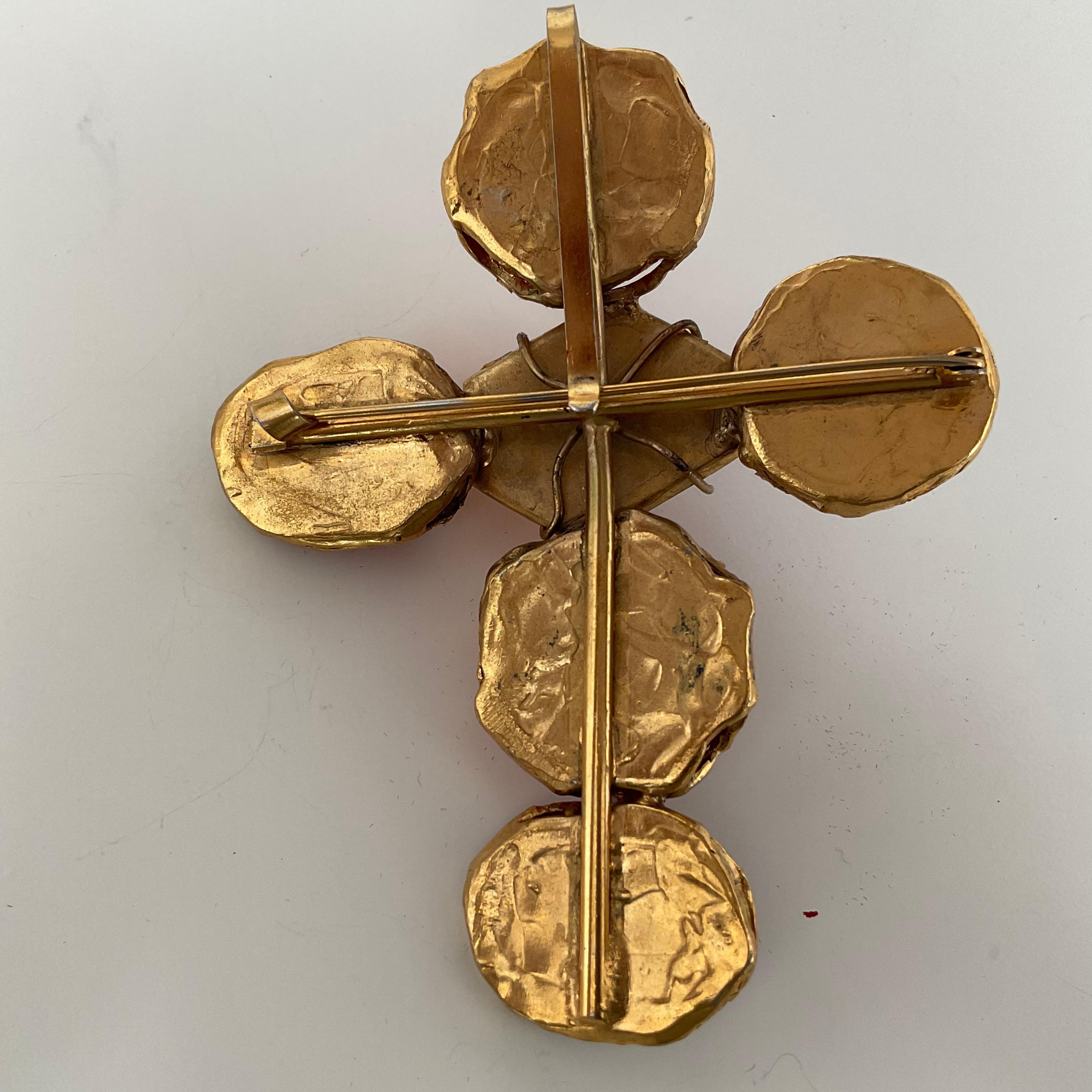 This singulare vintage piece from YVES SAINT LAURENT is a brooch from the 70's. It is designed as a cross with red and orange-toned stones in resin. A total of six stones are present, going from darker to lighter towards the centre. The orange stone