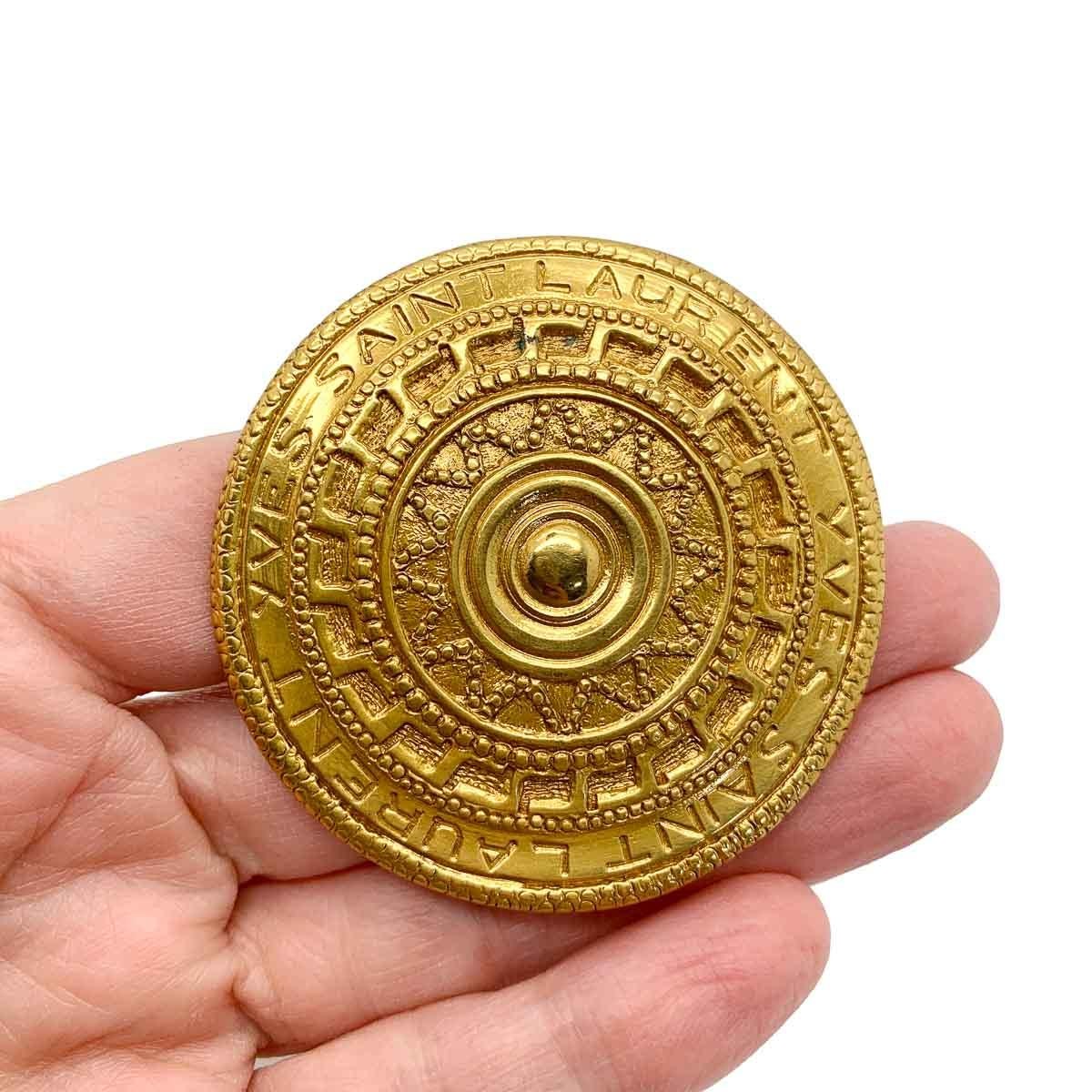 A wonderful vintage Yves Saint Laurent shield brooch. Featuring a large target style plaque embellished with engraving depicting concentric patterned circles and the name Yves Saint Laurent twice around edge.
In 1961, following his time as Dior's