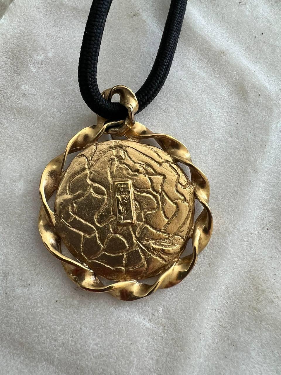 Vintage Yves Saint Laurent cord necklace with large medallion pendant. A thistle is engraved on the pendant made from gold-tone metal. 
Designed by Robert Goossens.  
Period: 1980s
Condition: very good. 
Dimensions:
Chain Length 25
