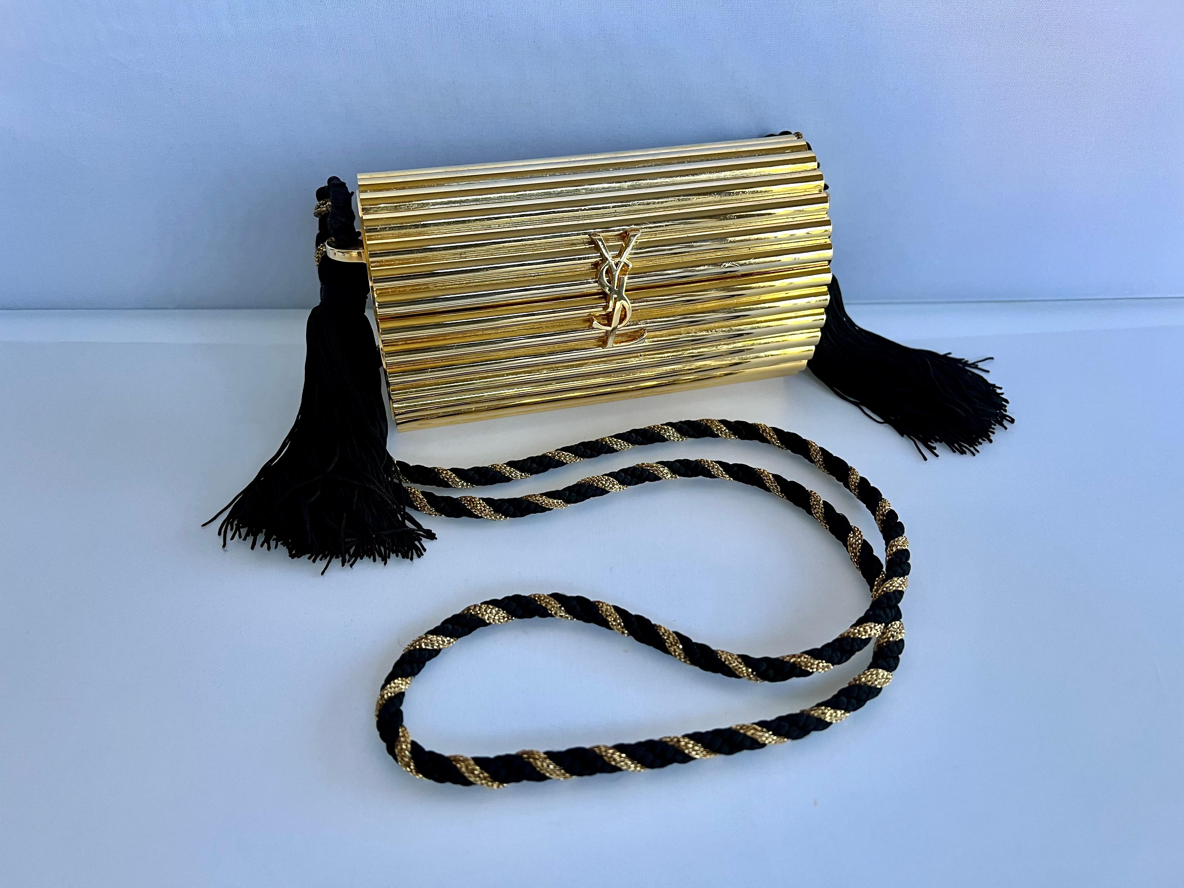 Highly collectable vintage Yves Saint Laurent (YSL) gold metal minaudire black tassel shoulder bag. Featuring -

- 100% Authentic YVES SAINT LAURENT.
- Gold plated ribbed rigid metal minaudière.
- Huge YSL clasp closure at the centre front.
- Strap