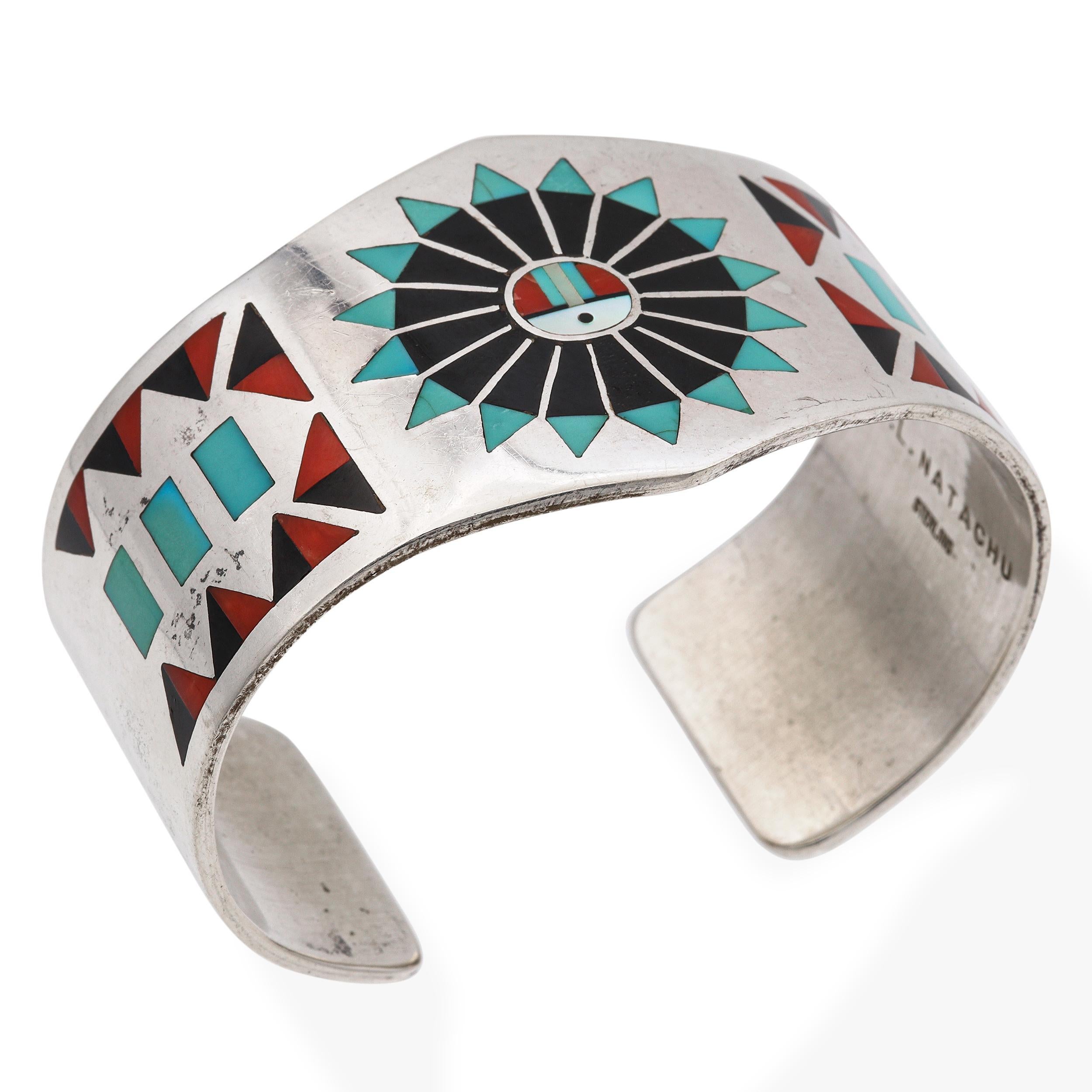 Un audaz ejemplo de artesanía Zuni, esta pulsera brazalete vintage está firmada por el artesano F.L. Natachu. La ancha banda de plata de ley está decorada con un intrincado trabajo de incrustación de piedras, con turquesa, coral, azabache y nácar en