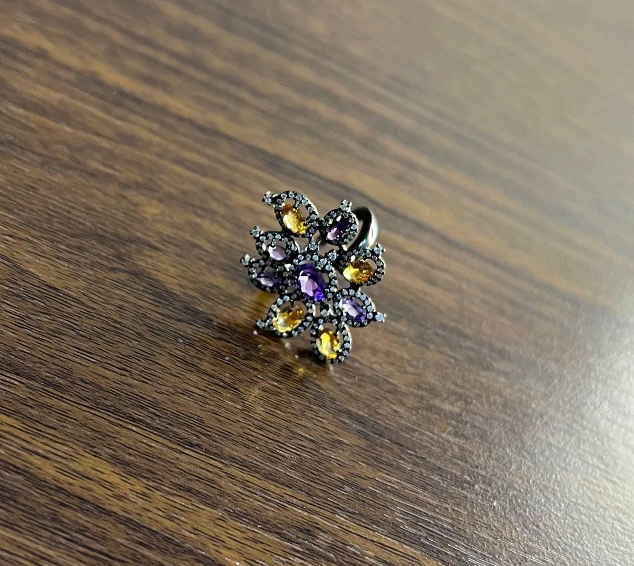 This striking floral ring captivates with its intricate design and vibrant gemstones.

Crafted in a14K Black Gold , it showcases a central oval amethyst surrounded by a halo of smaller round diamonds, creating a vivid focal point. Encircling the
