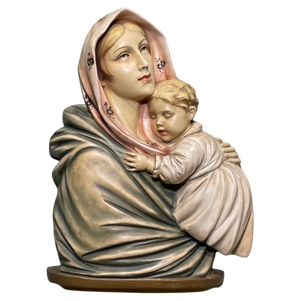 This a sculpture/relief of the Virgin Mary holding very tenderly a Baby Jesus who fell asleep. It is attached to an almost square wood frame. The sculpture stand over an oval wood base. Her eyes are blue and looking up with a serene gaze ( like