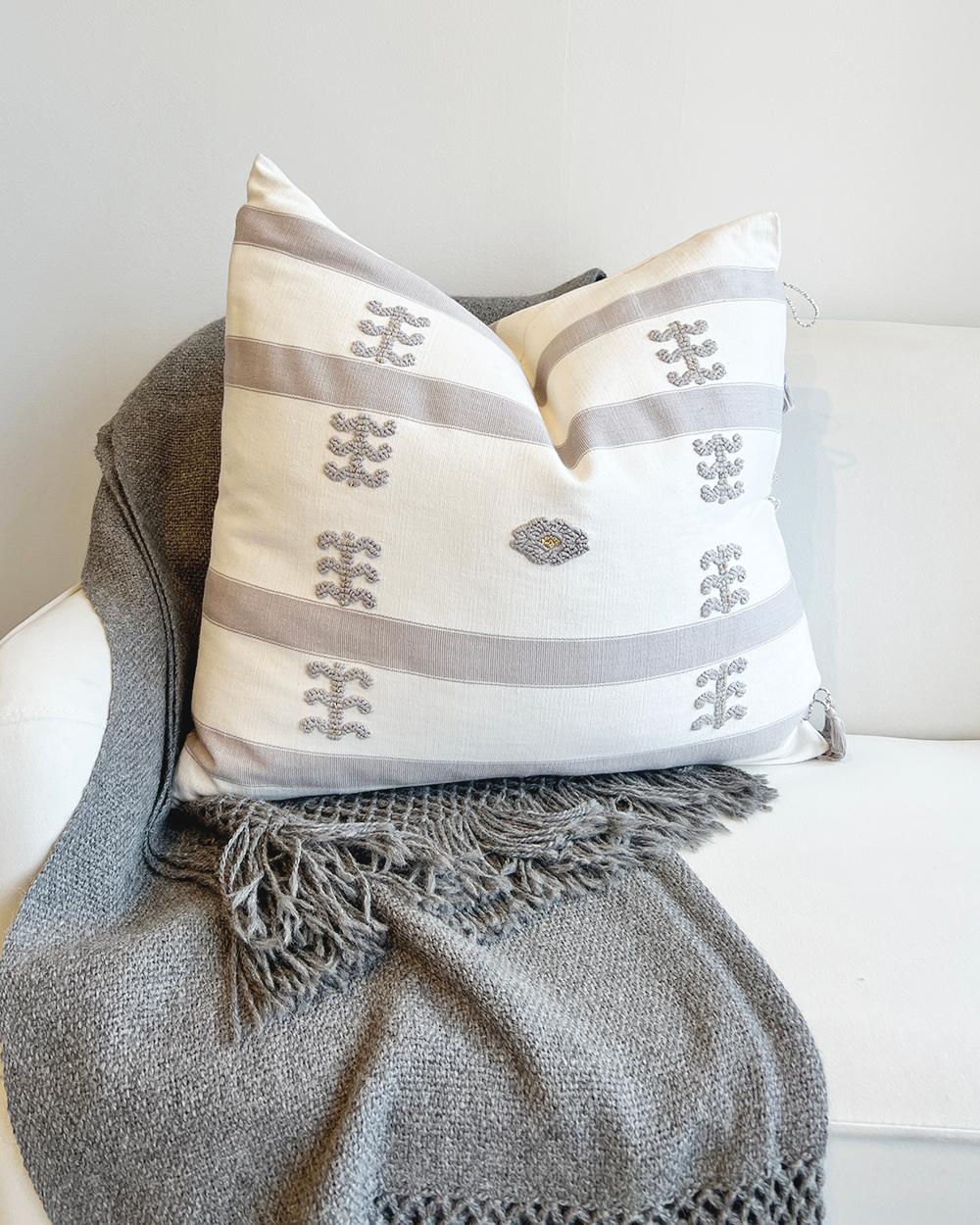 This handcrafted Mexican-inspired fair trade cotton throw pillow adds style and comfort to any space. The minimalist design features a white background with elegant gray embroidery and rustic accents, evoking a modern and cozy atmosphere. Add a
