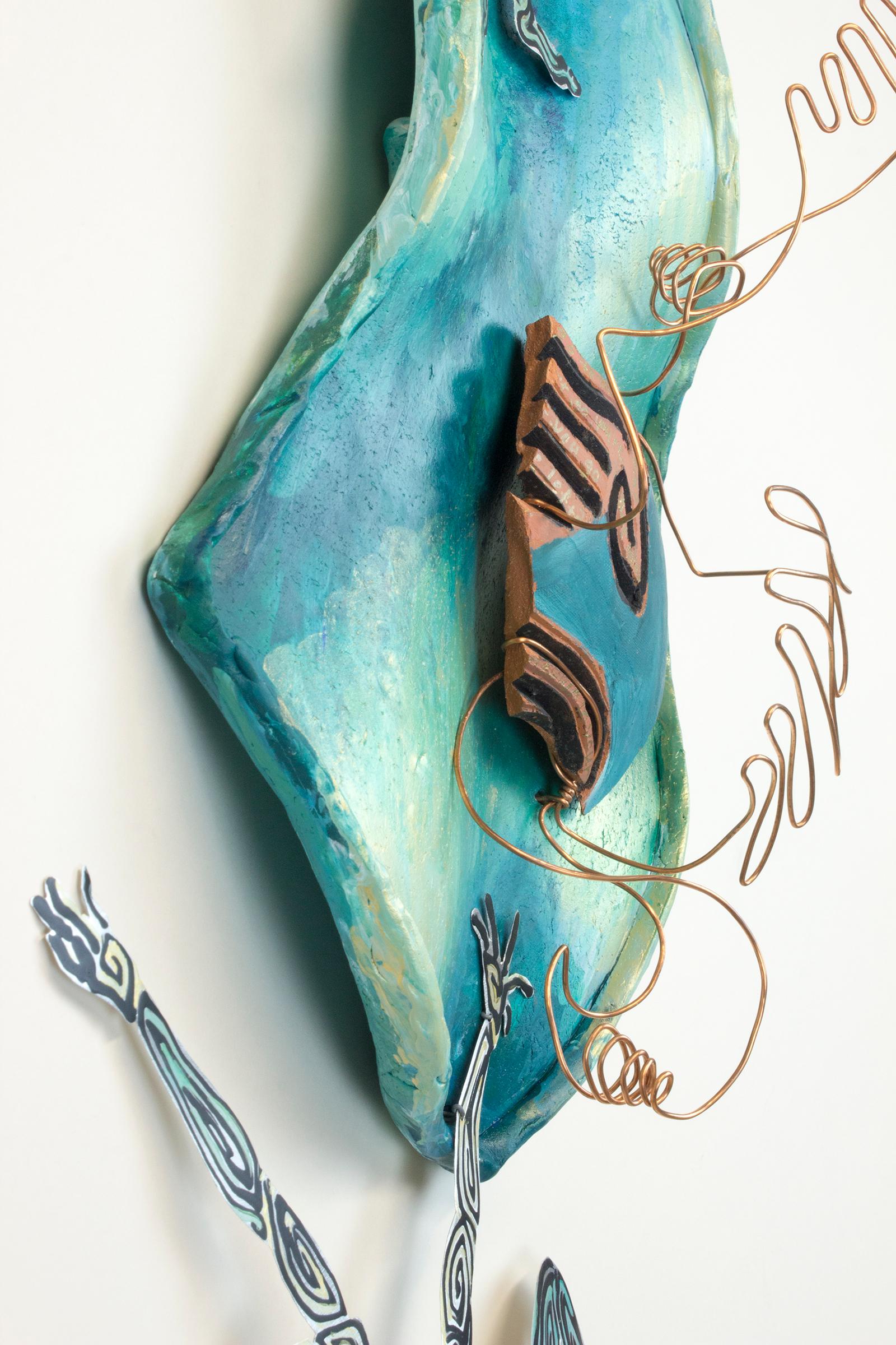 Virginia Mahoney’s “Unresolved Contradiction” is a 25 x 13 x 6 inch ceramic and metal wall sculpture in teal, gold, blue, black, silver, and brown. Two cutout figures in silver, blue, and black painted metal interact playfully with a ceramic shard