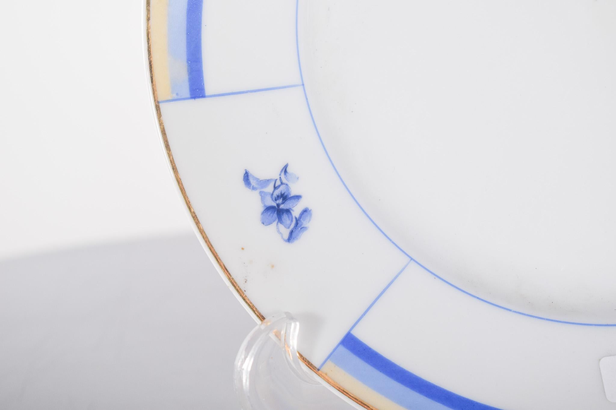 Vista Alegre porcelain plate. Minimalist decoration in blue and gold on a white background.
The rim of the plate is decorated with geometric patterns made up of blue and gold bands, interspersed with small hand-painted floral motifs in blue, giving