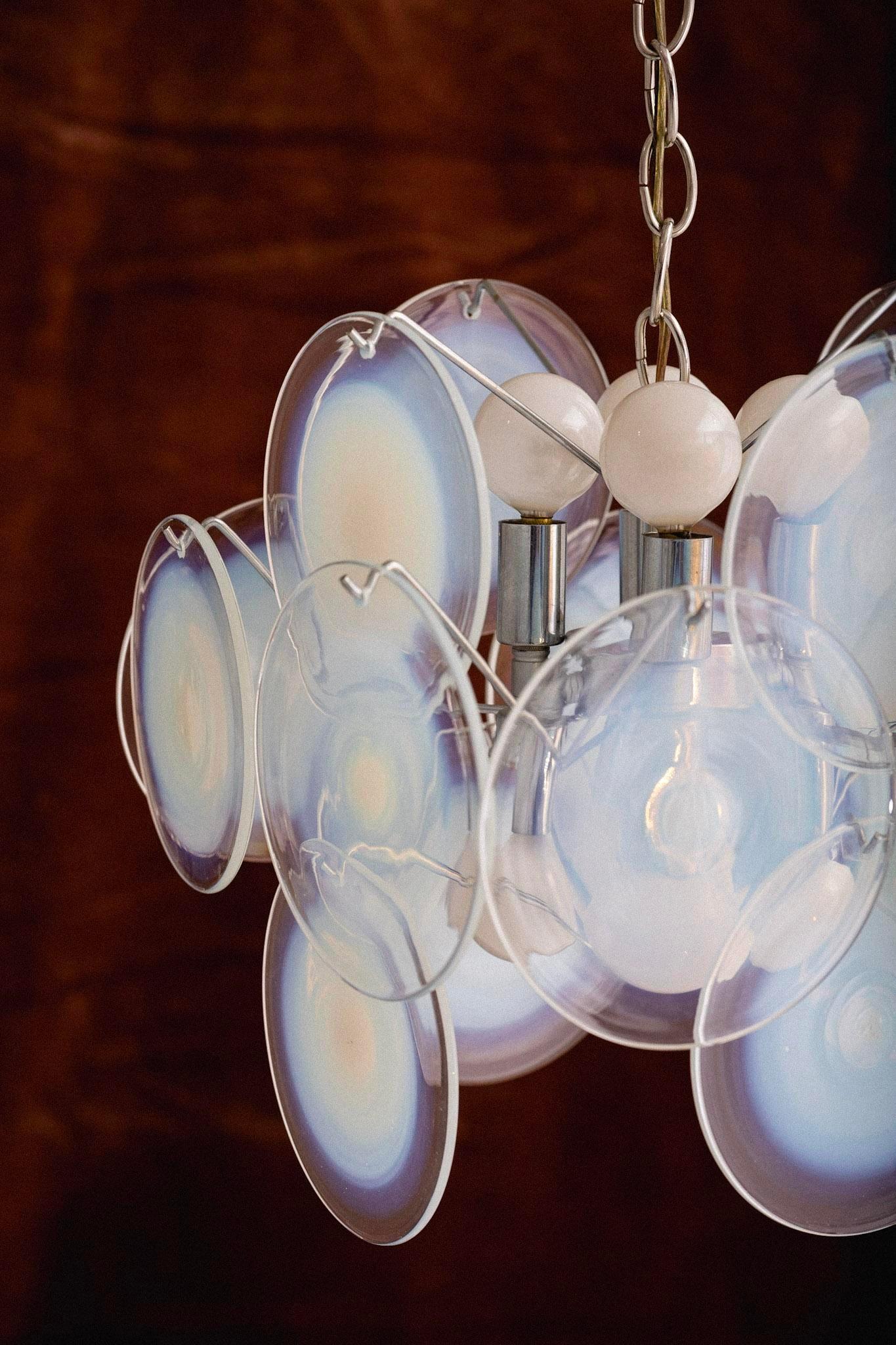 A glass chandelier by Vistosi Murano. 16 glass discs hang on a chrome frame. Each hand blown glass disc ranges from clear glass to an opaque opalescent center. Each disc measures 6” in diameter. The chrome frame measures 14” wide by 14” deep by 10”