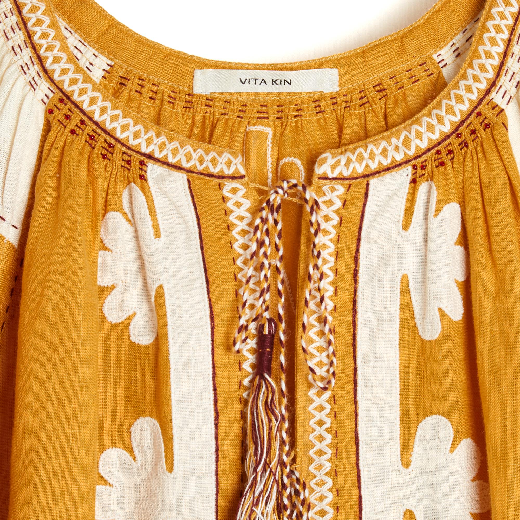 Vita Kin dress, Shalimar model, in saffron yellow linen embroidered with large ethnic-inspired patterns (Ukraine) in ecru and white, round slit collar closed by 4 (2x2) ties and multicolored pompoms, long puffed sleeves closed by a button, 2 slit