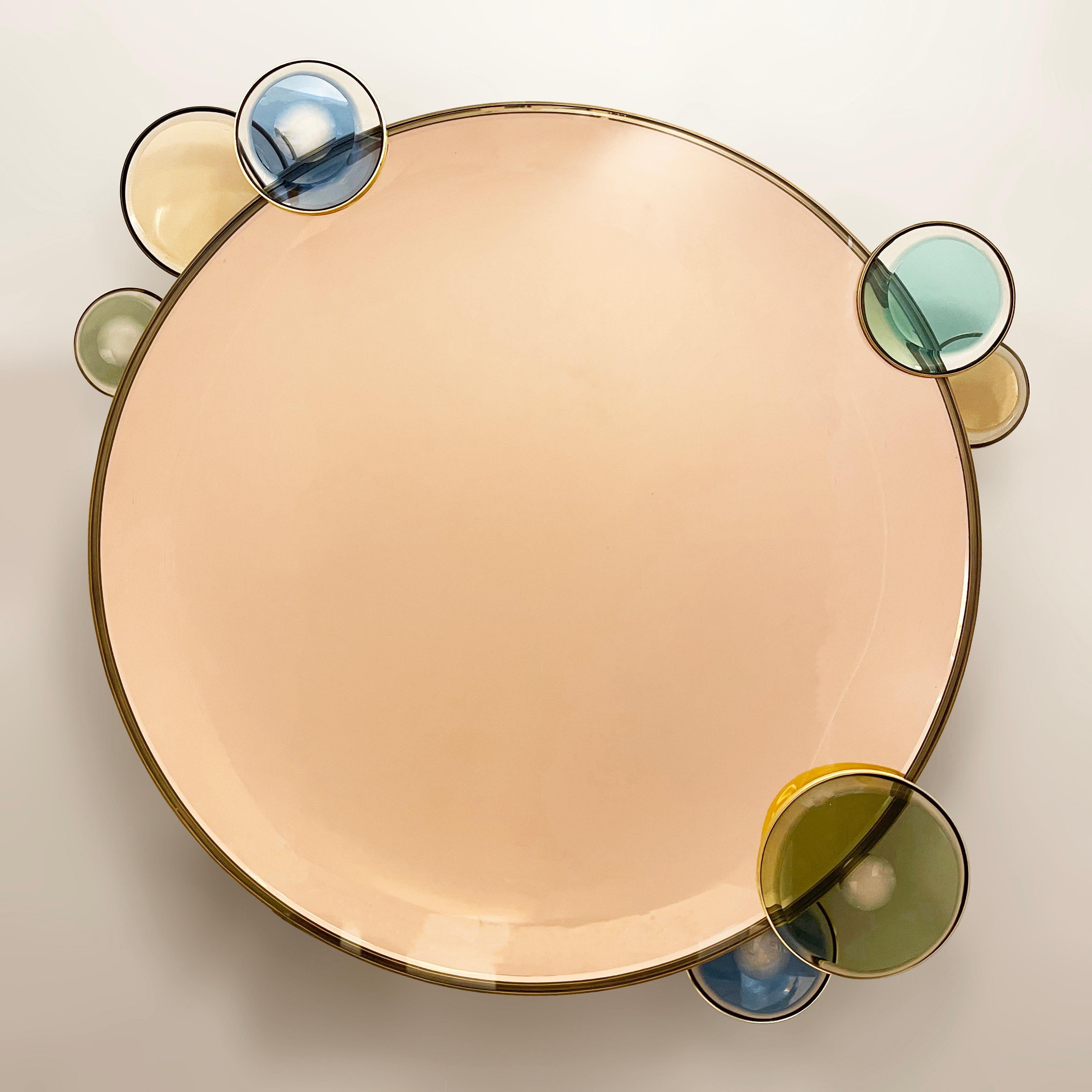 The Vitreus with its tinted reflecting concave glass, lends itself to be both mirror and wall sculpture. Colored glass medallions mounted to its brass frame provide an additional pop of color while a flat clear glass on top of the concave mirror