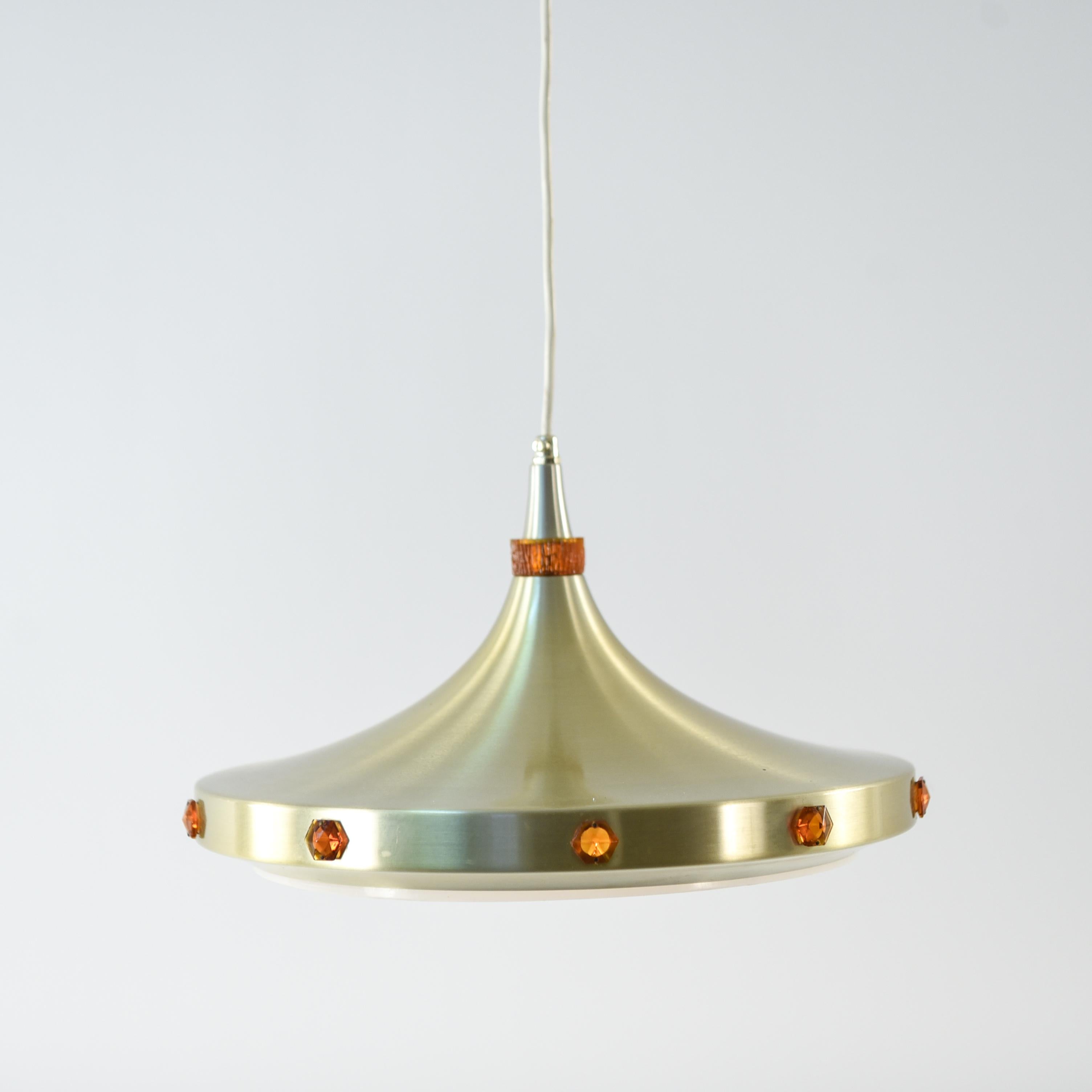 This Danish midcentury pendant light by Vitrika features a brass outer ring adorned with orange colored glass jewels. On the interior are white concentric circles which not only function to reflect more light, but also add an element of stylistic