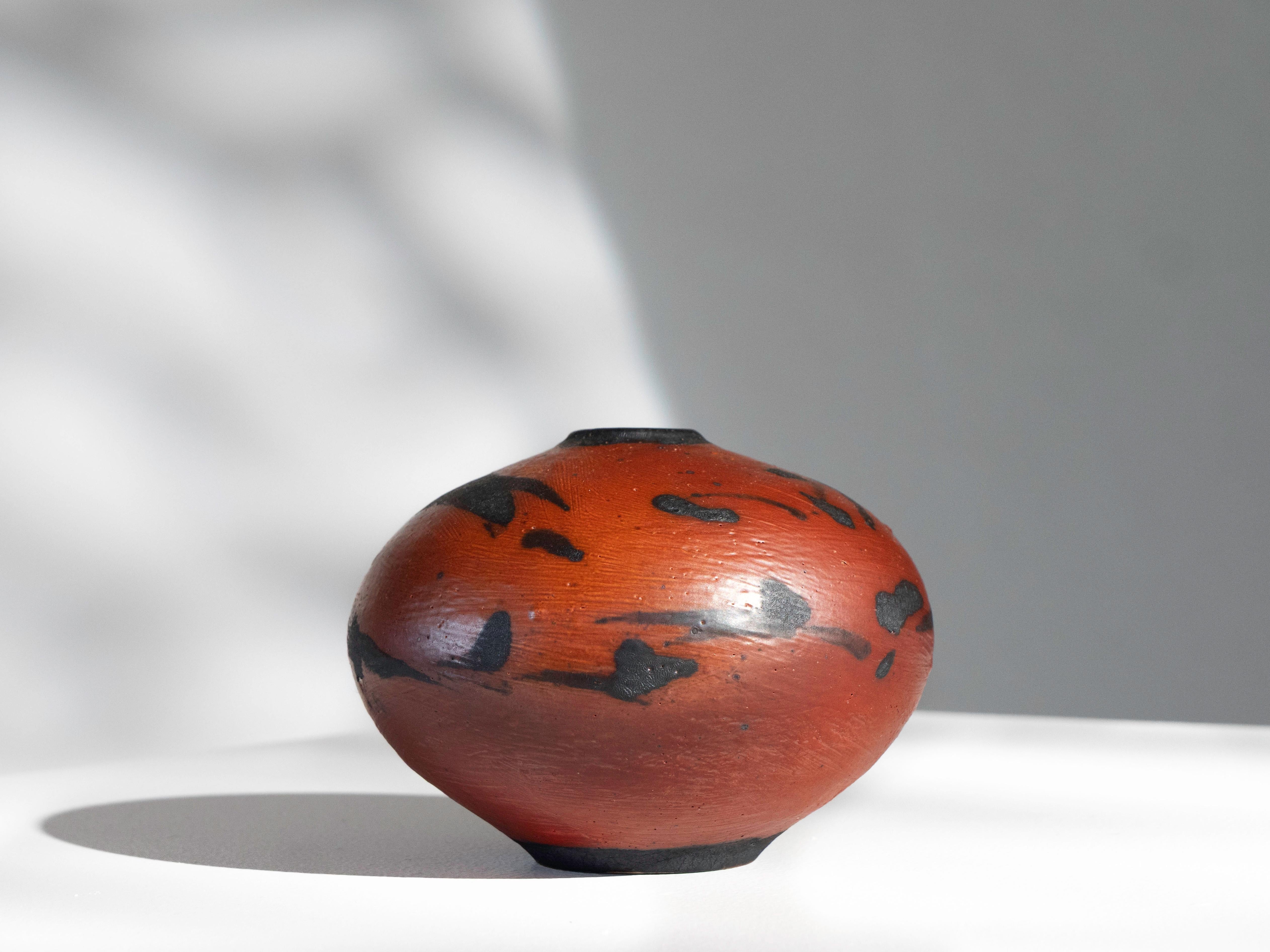 Petit récipient en poterie de studio du céramiste Otto Heino. Ce vase en forme de bourgeon arrondi présente une base aux tons rouges et orange ornée d'une texture de type craquelure noire. La texture de la finition montre des coups de pinceau autour