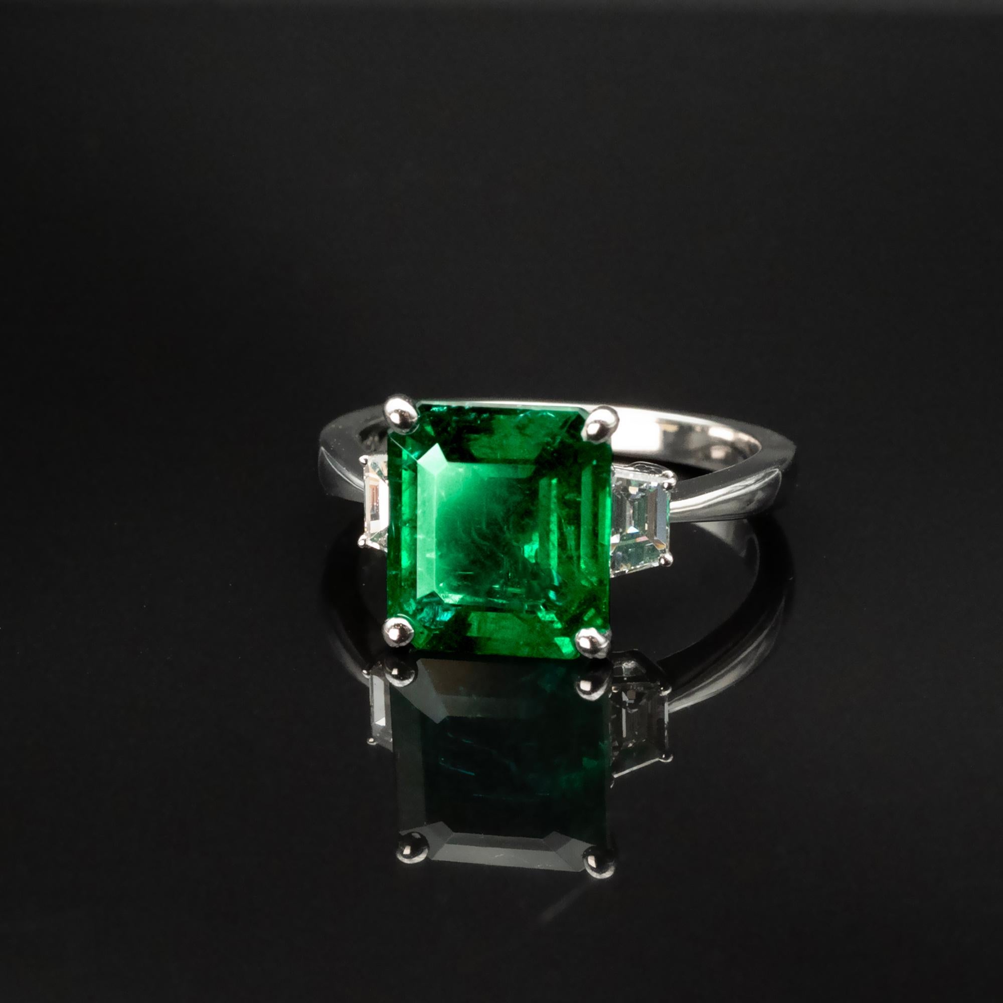 Introducing a stunning 18-karat white gold emerald three-stone ring. 
This exquisite piece features a magnificent center natural emerald weighing 2.98 carats, a true marvel of nature. The gemstone is certified by Bellerophon Gemlab and boasts a rich