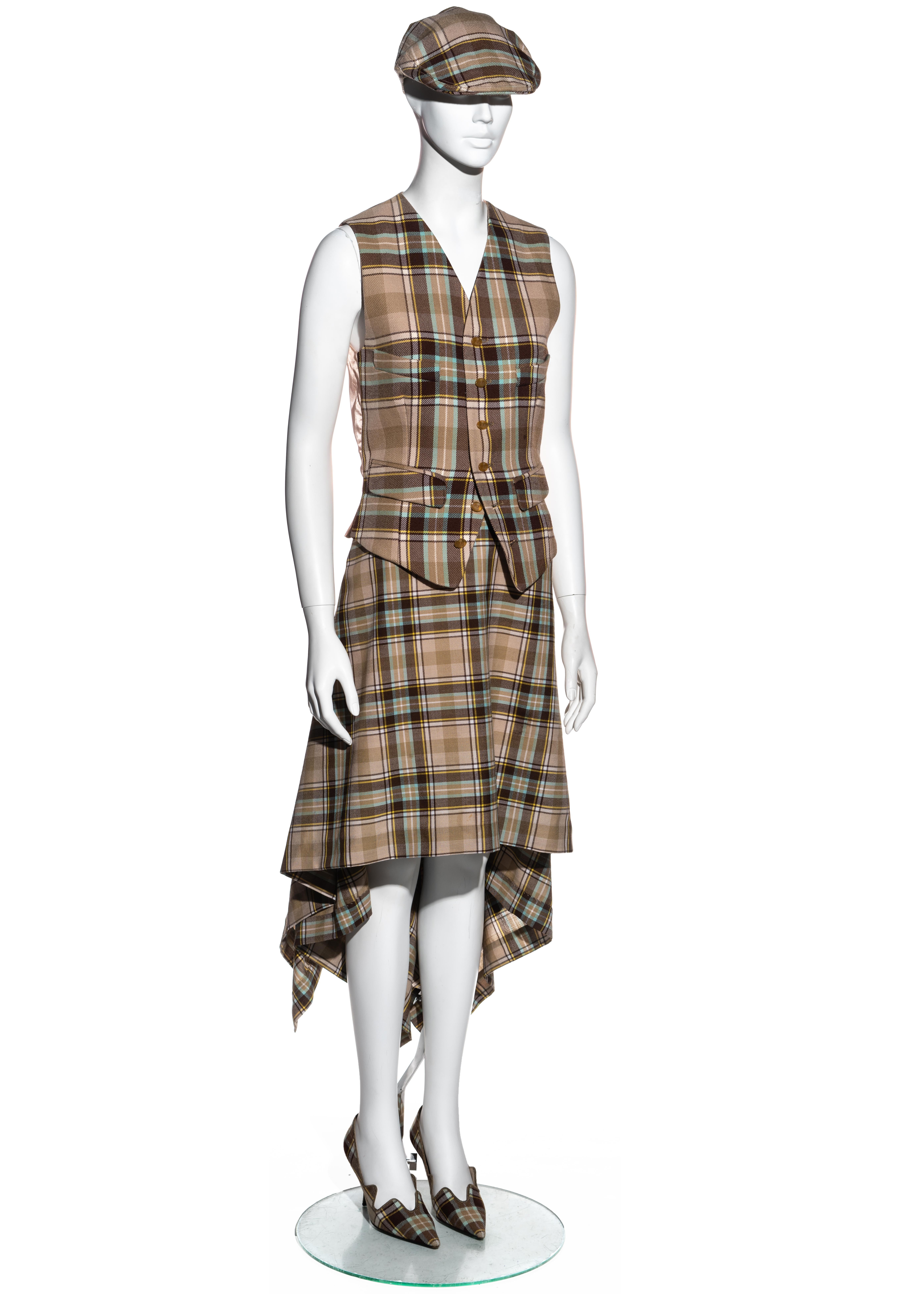 ▪ Vivienne Westwood beige tartan wool four piece ensemble 
▪ Beige, brown, yellow, white and blue tartan wool 
▪ Waistcoat with orb engraved buttons and pin satin back panel 
▪ Mid-length wrap skirt with descending hemline 
▪ Flat-cap with