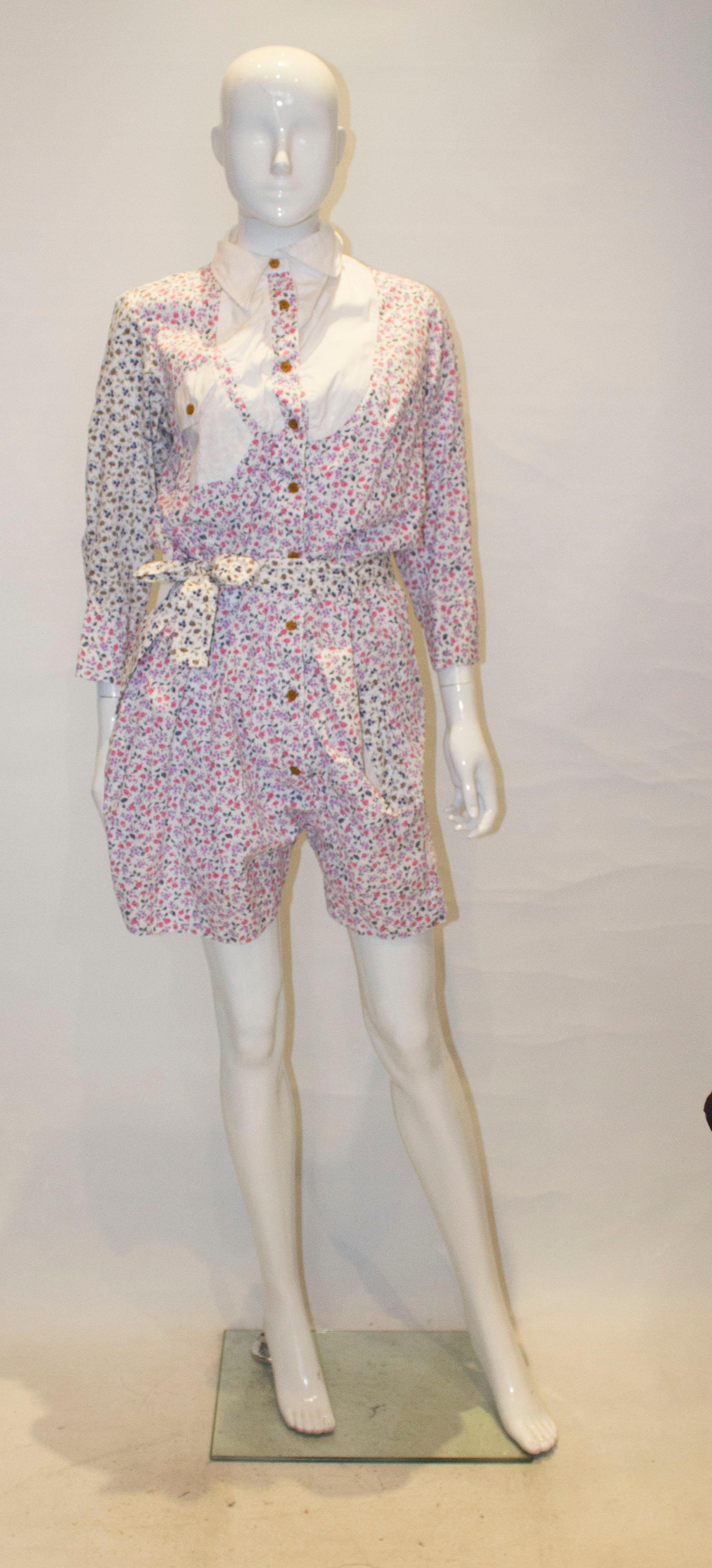 A fun playsuit in floral cotton by Vivienne Westwood, Red Label. The suit has a white collar, yoke and pocket, with a nine button front opening. It has a self fabric belt and pockets front and back.