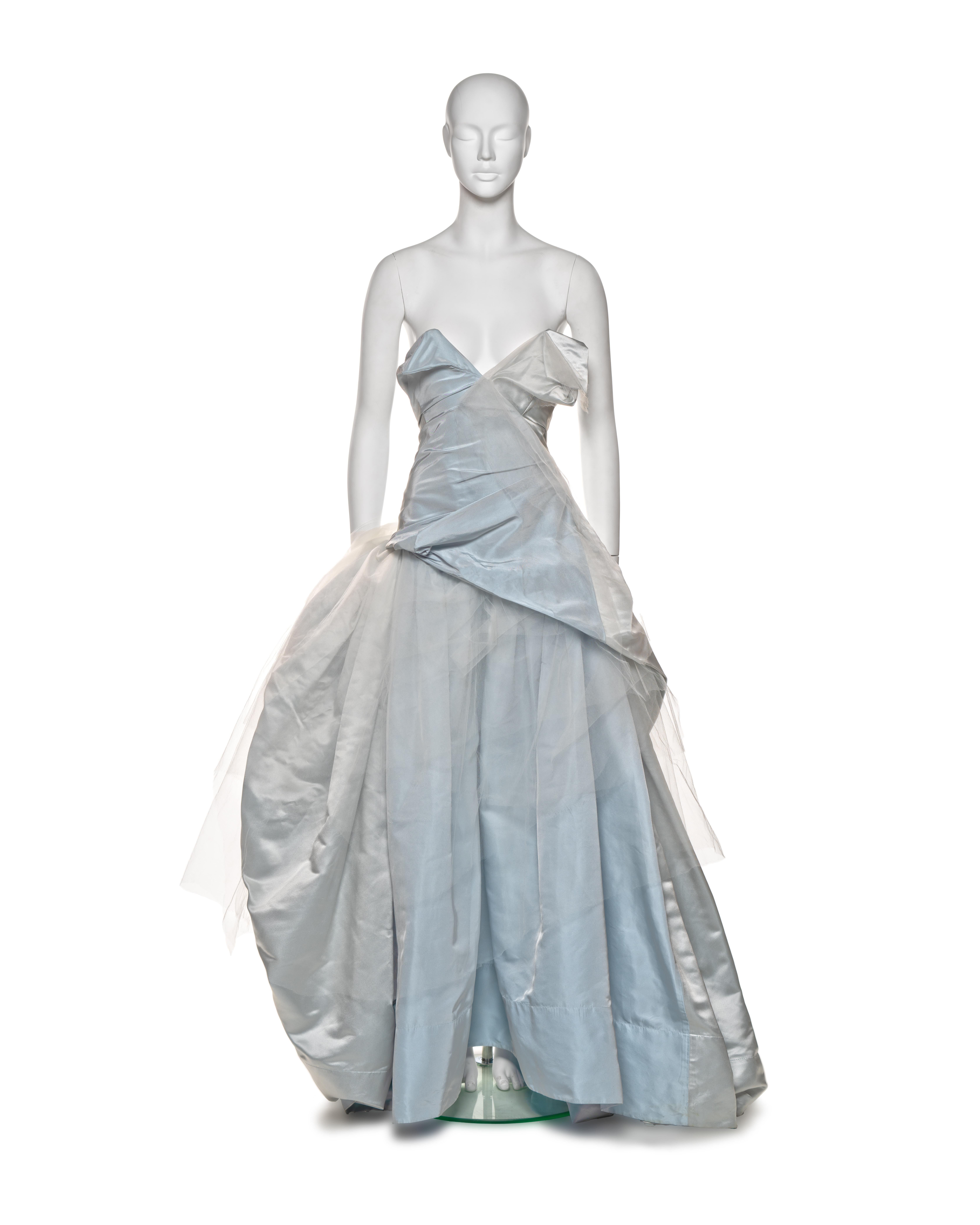 This Vivienne Westwood bridal couture dress is crafted from pale blue silk taffeta, featuring an asymmetrical bodice design with a built-in corset for structure. The dress has a voluminous skirt, enhanced by layers of tulle and silk, which can be