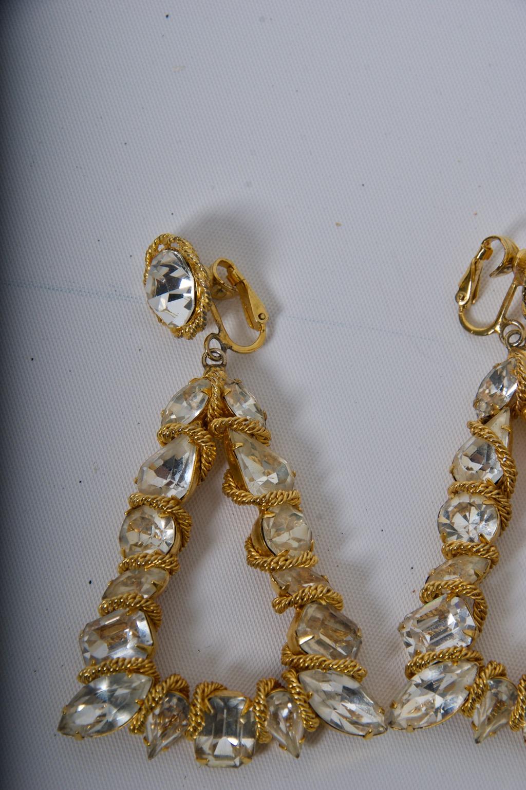 Grandi e scenografici orecchini di Vogue Jewelry di forma triangolare e composti da grandi cristalli rotondi e marquise in un'incastonatura dorata, ogni pietra intrecciata con una sottile catena continua. I triangoli sono sospesi da un singolo