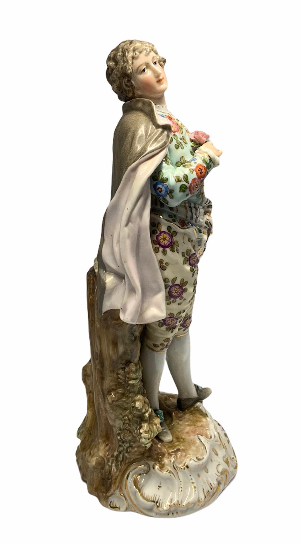 This is a Volksted Porcelain depicting a gentleman dressed with two different colorful flower fabrics in his blouse & pants. His shoes are adorned with flowers. He is holding a rose in his right hand & a gentleman cane in his left hand. A grey cape