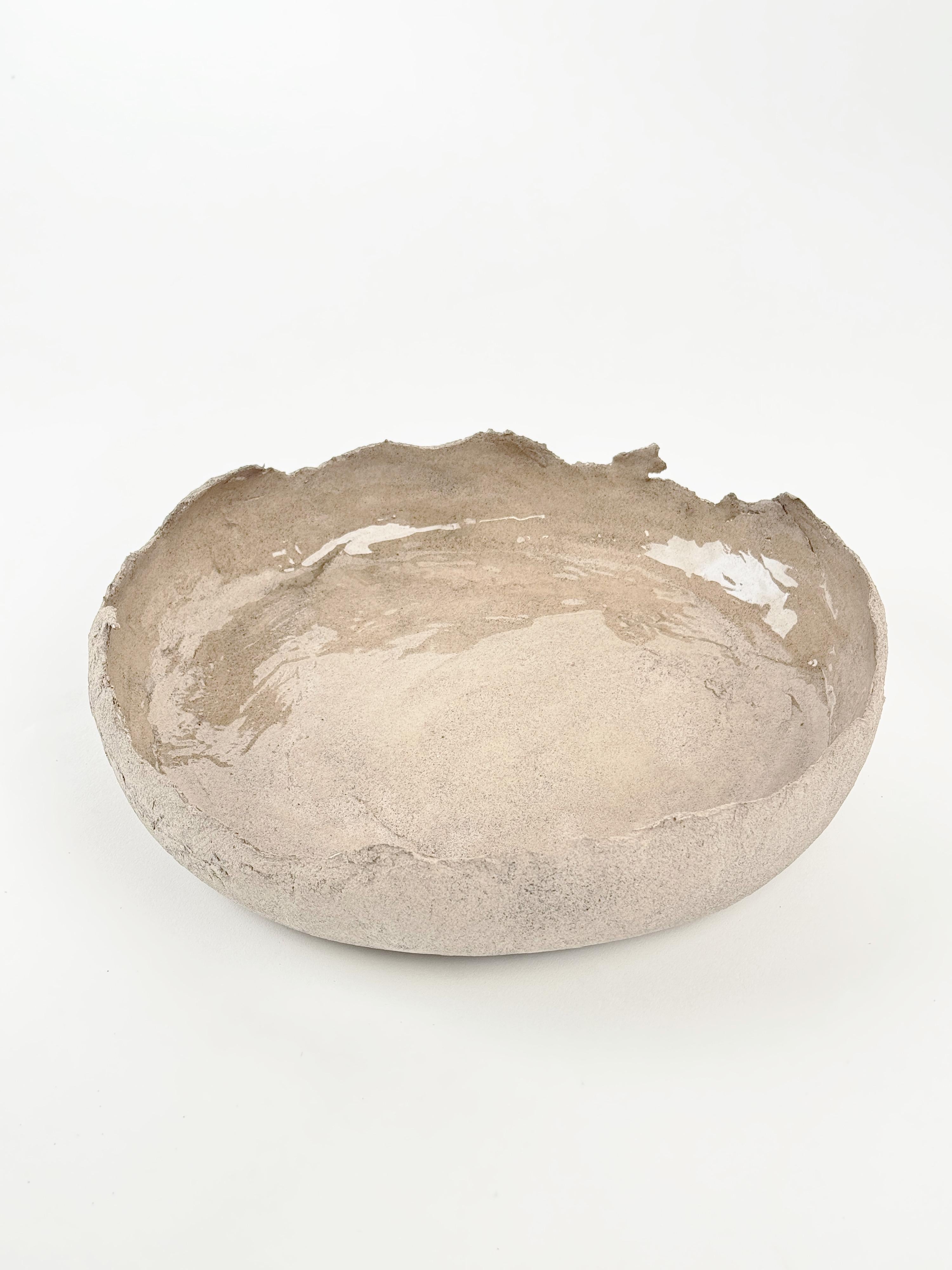 Este cuenco escultórico de cerámica es una pieza única, hecha a mano con gres natural. Su forma baja y ancha se complementa con un borde desgarrado orgánicamente y una rica textura superficial, que revela capas de tierra cruda y refinada artesanía.