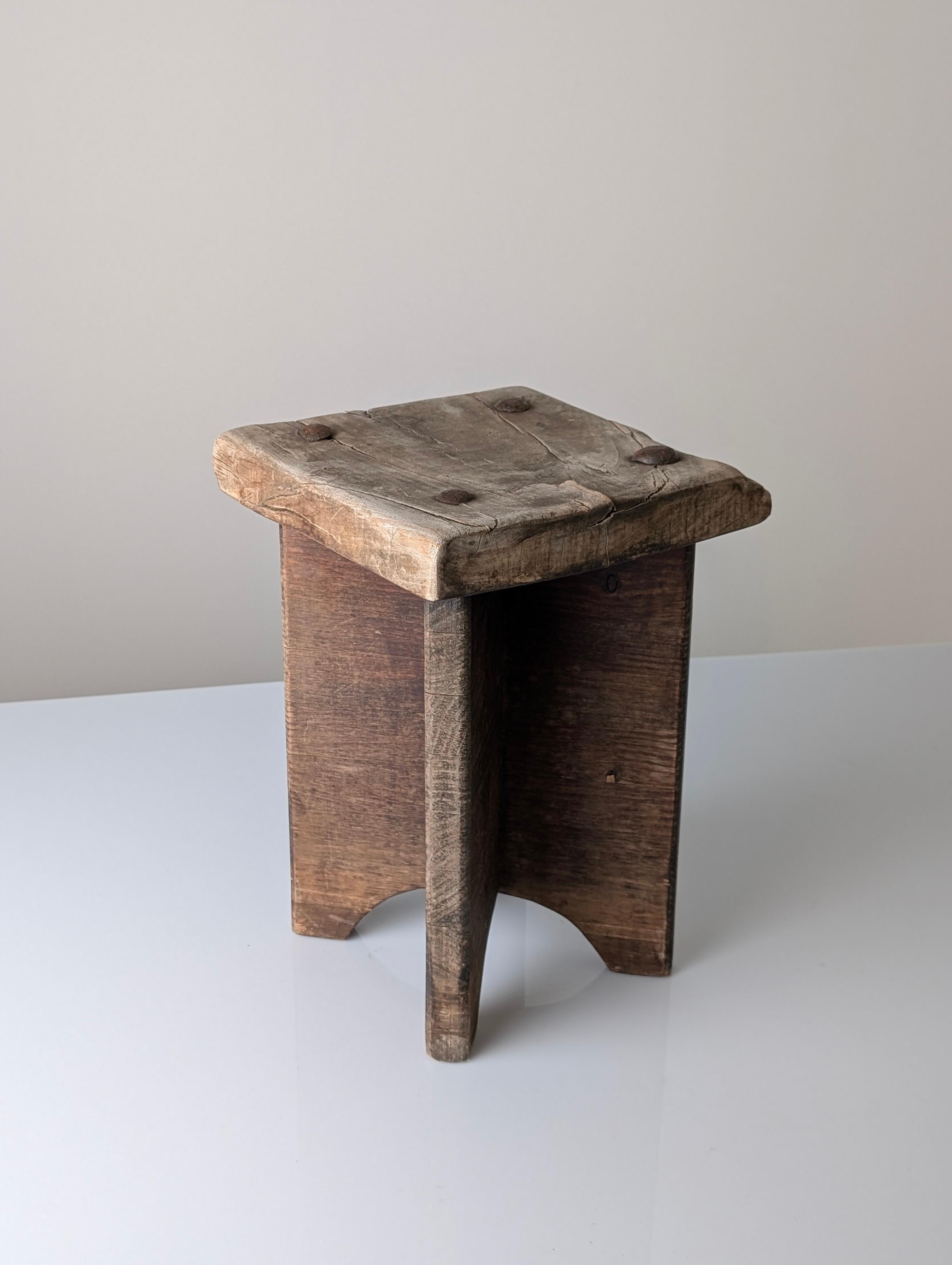 Sgabello rustico in legno del 1800, che riflette la filosofia wabi sabi con le sue irregolarità naturali, l'usura visibile e la patina invecchiata. La seduta quadrata presenta profonde crepe e chiodi forgiati a mano, mentre la semplice struttura a