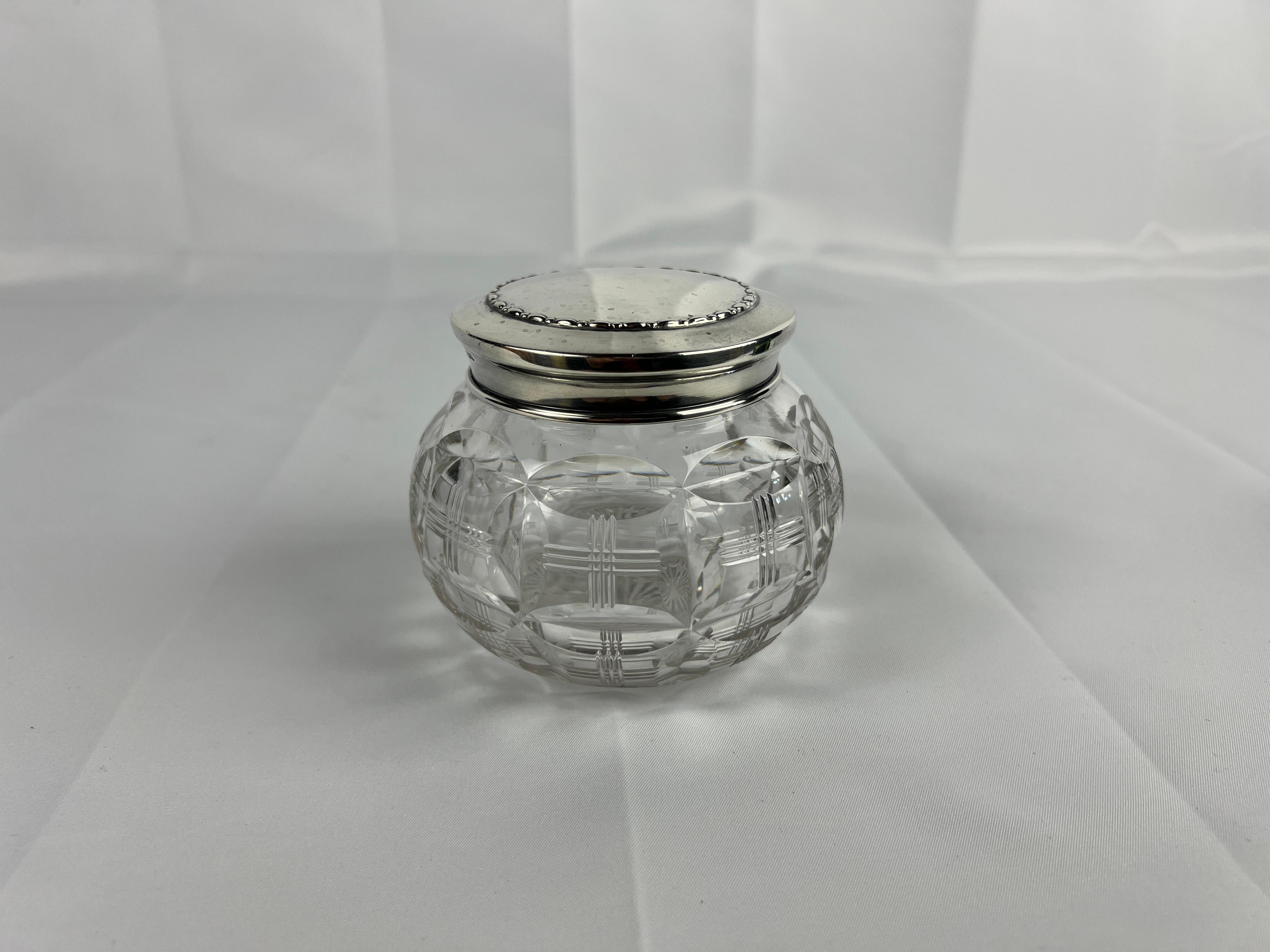 This elegant vanity jar by Walker & Hall of Sheffield exemplifies the sophistication of early 20th-century British craftsmanship. The jar features a beautifully cut glass body, showcasing intricate patterns that catch and refract light, creating a