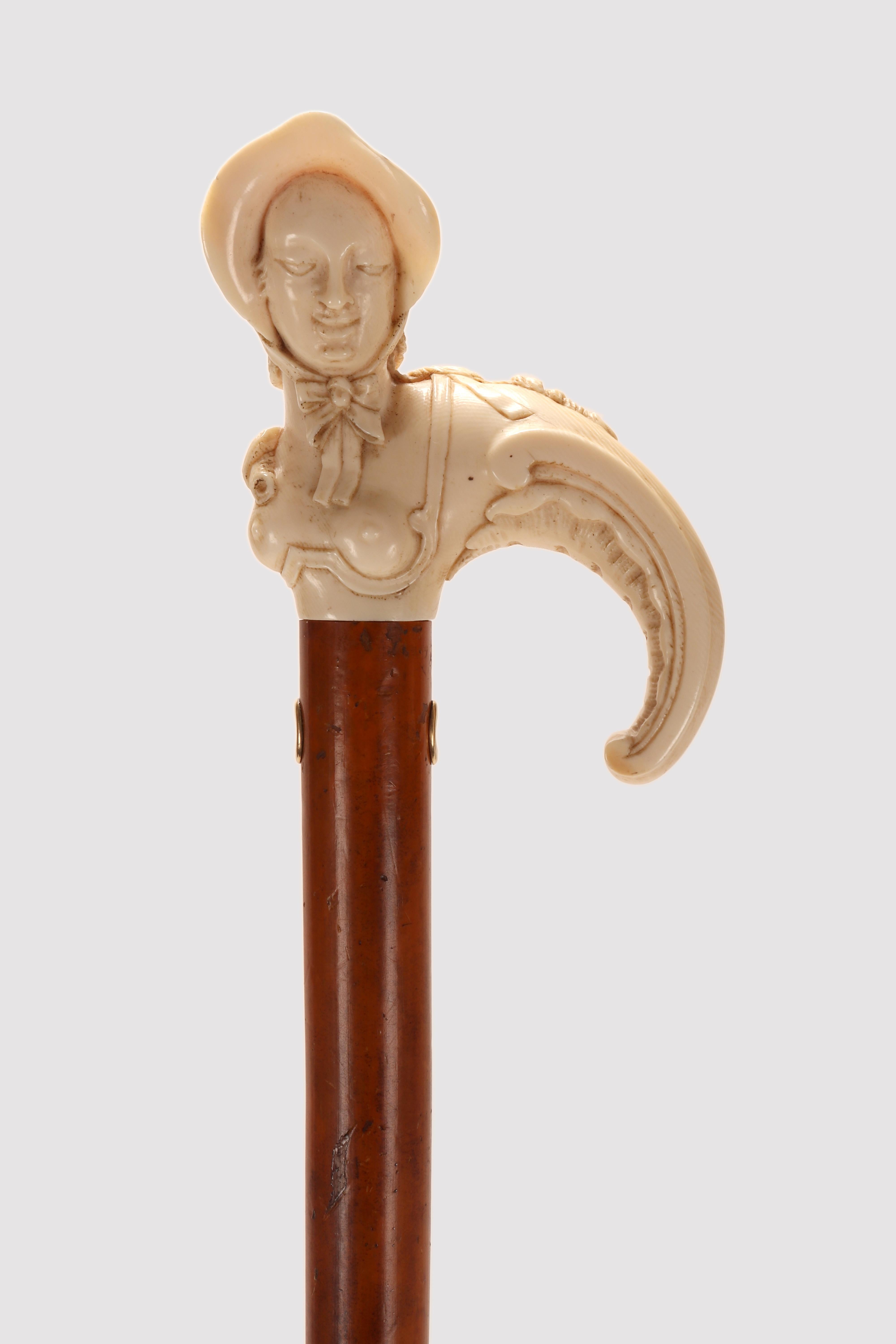 Bastón: Gran mango de marfil finamente tallado en forma de bequille. Representa un busto femenino, una mujer de pechos desnudos que lleva un sombrero de paja finamente detallado. Esta forma típica de asa, con volutas que terminan en una voluta, se