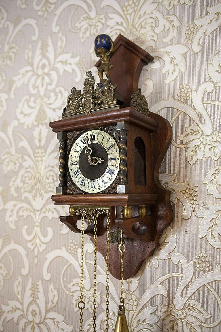 Wall Clock From the Late 20th century in Oak Case

The case is wooden, with glazed sides. Above the clock face, there is a brass crown.
The figurine of Atlas carrying the globe can be seen in the upper section. Furthermore, the pendulum is shaped