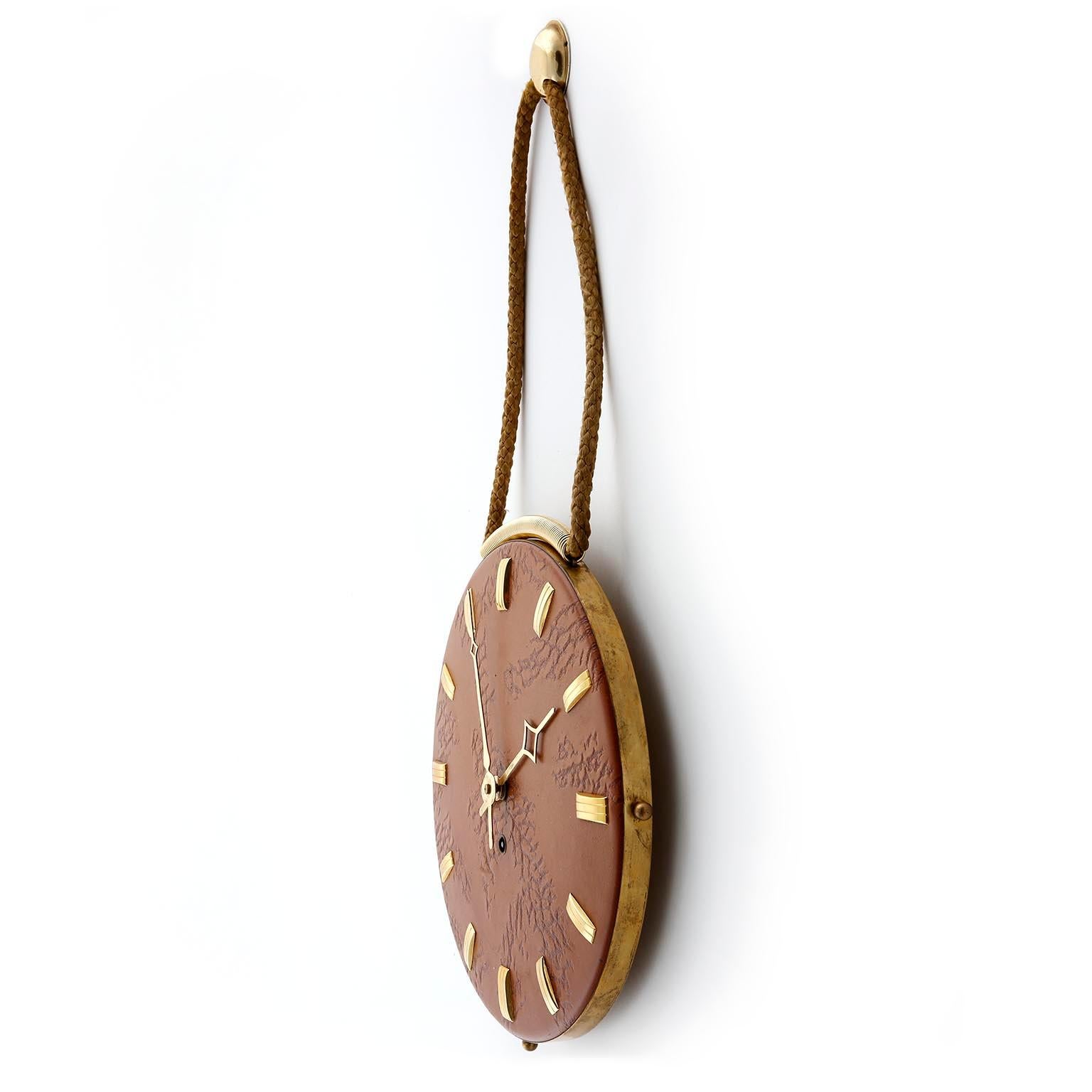 A wall clock made of leather and brass manufactured in midcentury, circa 1960.
The clock hangs with golden toned cords on a brass knob.
A removable key is used for winding the movement.
The clock is very good condition and in working