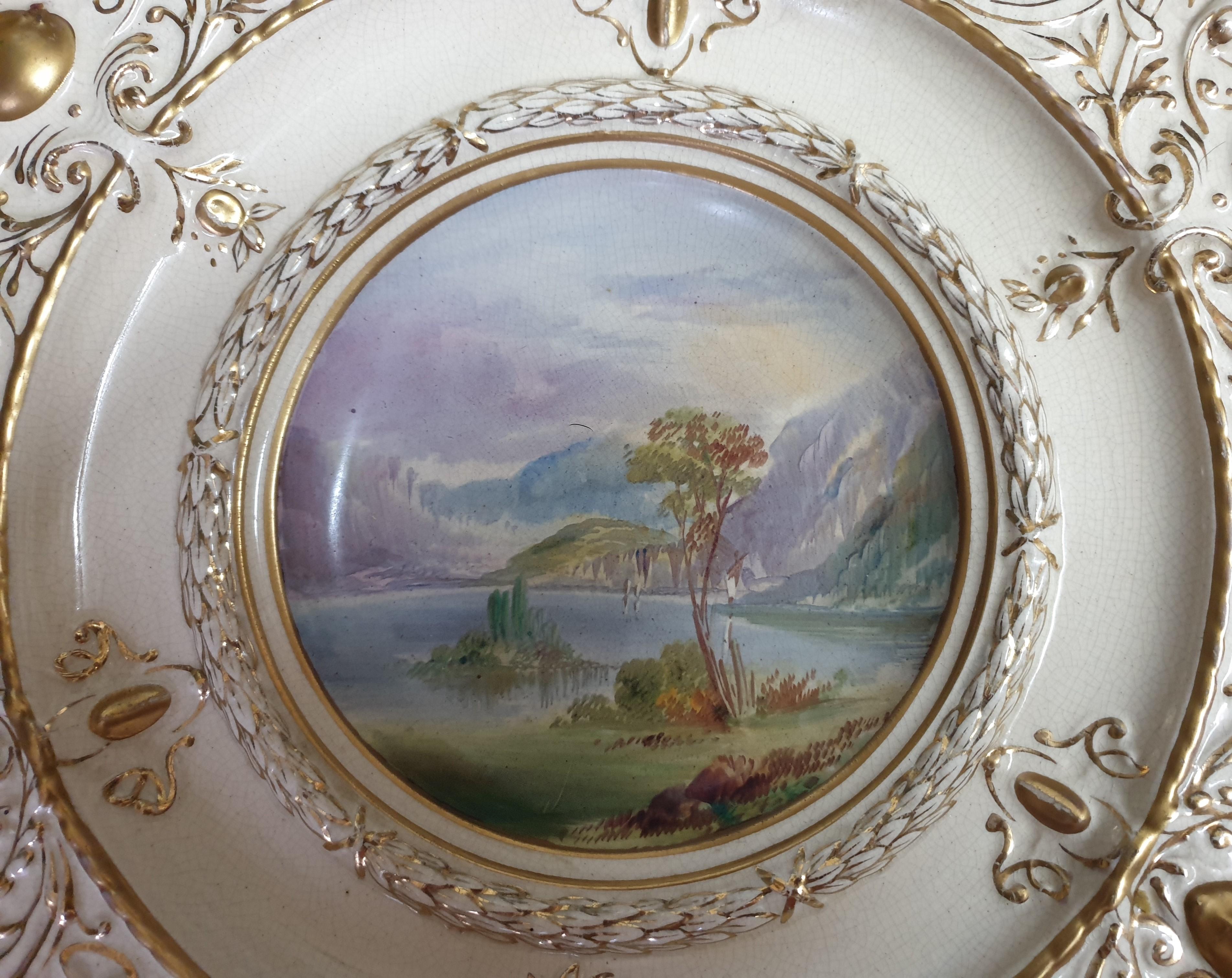 A large Wall decorative charger depicting a lakeside scene with a mountainous backdrop. The scene is handpainted on transfer a technique used to make handpainted porcelain more affordable. The rim is highly decorated with gilt featuring scarabs,