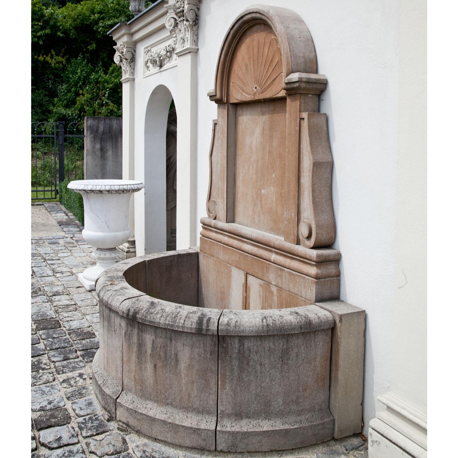 Wall fountain out of hand-carved patinated sandstone with a semicircular basin (H: 62 cm) and a tall rear wall with a relief shell décor, a rounded pediment and volute details towards the sides. The spout is installed in the center of the shell