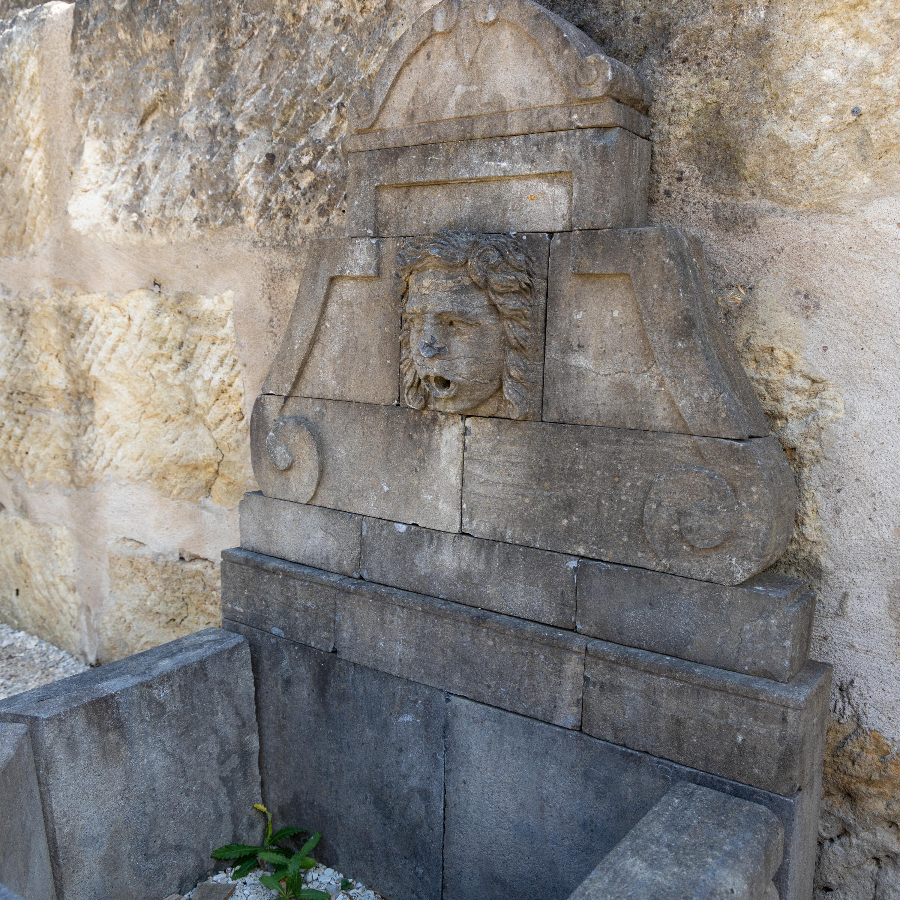 Wandbrunnen mit sechseckigem Beckenrand und hoher Rückwand mit Volutendekor und zentralem Maskaron als Wasserspeier. Blaustein, handgeschnitzt.