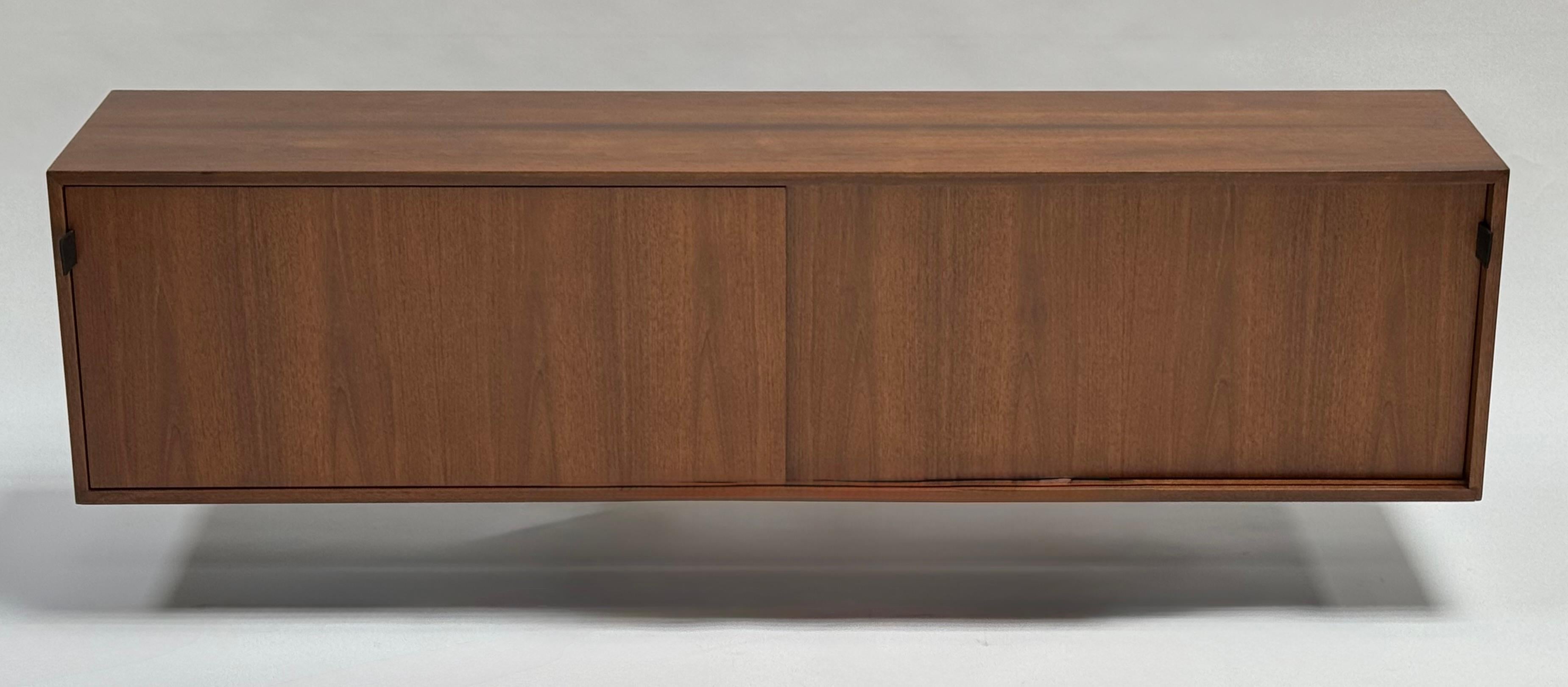 Wall hanging credenza designed by Florence Knoll and produced from 1947 to 1973, this is a 1960s piece. Walnut top, sides and doors with black leather pulls. The interior is a lacquered off white with four bays, two with adjustable oak shelves. The