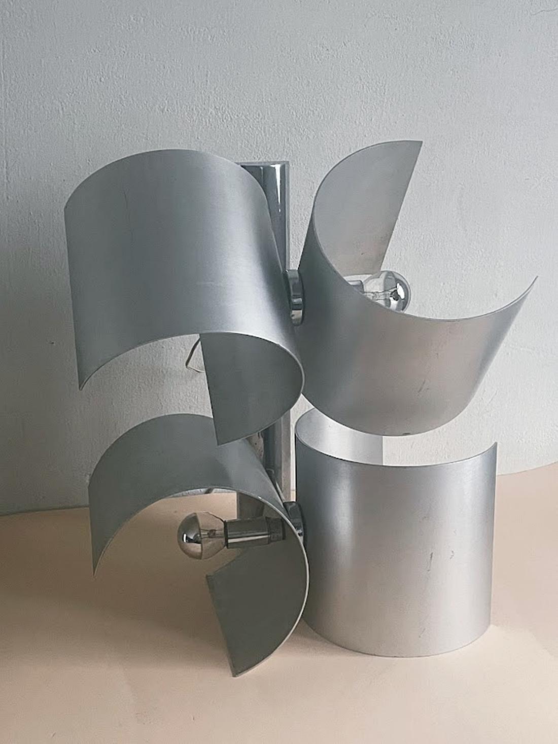 El aplique, fabricado por Nucleo Sormani en la década de 1970, consta de cuatro piezas curvadas de aluminio que giran independientemente, cada una de ellas equipada con una toma E14. Este diseño permite personalizar la dirección y distribución de la