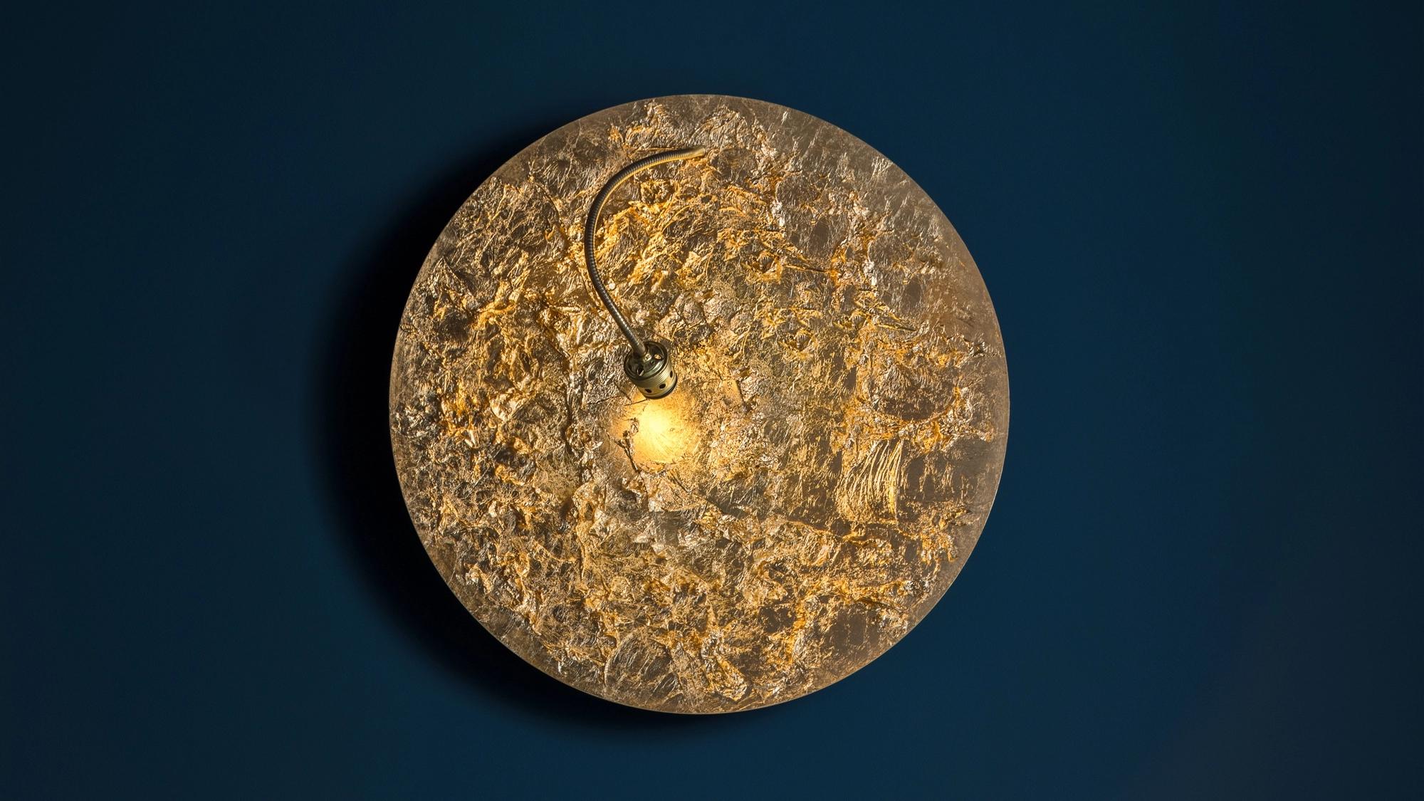La Luna Piena, de la colección Luci d'Oro de Enzo Catellani, es una lámpara de techo o de pared que celebra la luz dorada del sol y del fuego. Su disco de aluminio de mayor tamaño, revestido de hojas voladoras, crea un resplandor llamativo y cálido