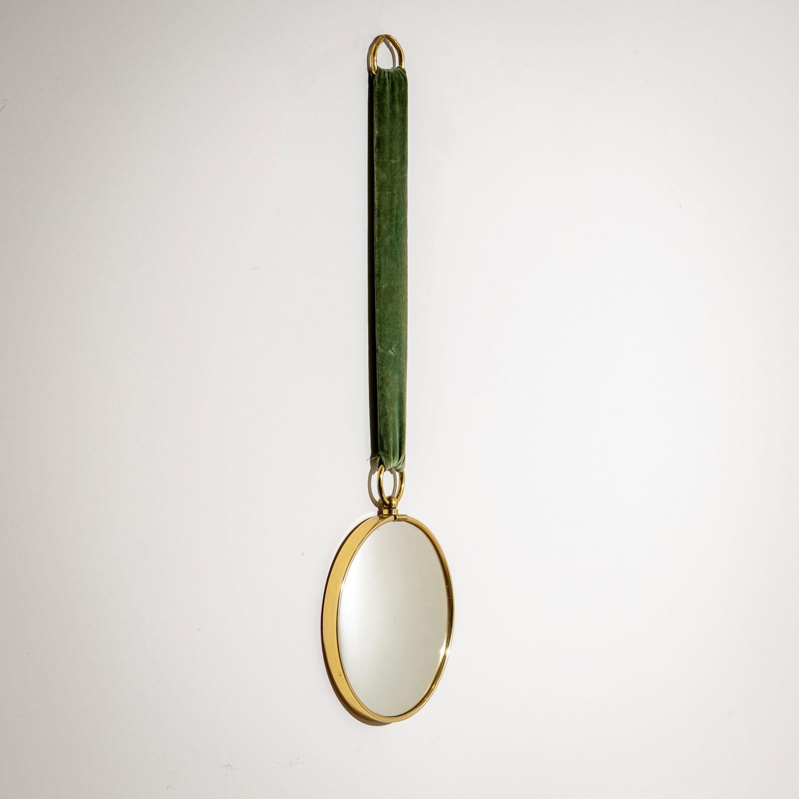 A round wall mirror with a brass frame, featuring a large ring that gives it the appearance of an oversized pocket watch. The mirror is suspended by a green velvet ribbon, adding a playful and imaginative character. A charming and stylish accessory