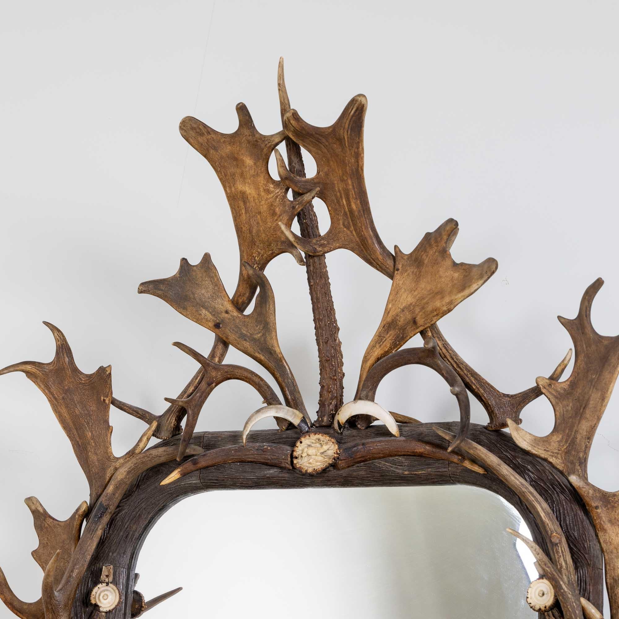 Large rectangular wall mirror with a large frame with rounded corners and symmetrical decoration of deer antlers and tusks. The ends of the antlers are partly decorated with small carvings in the shape of deer. Smaller decorative parts missing.