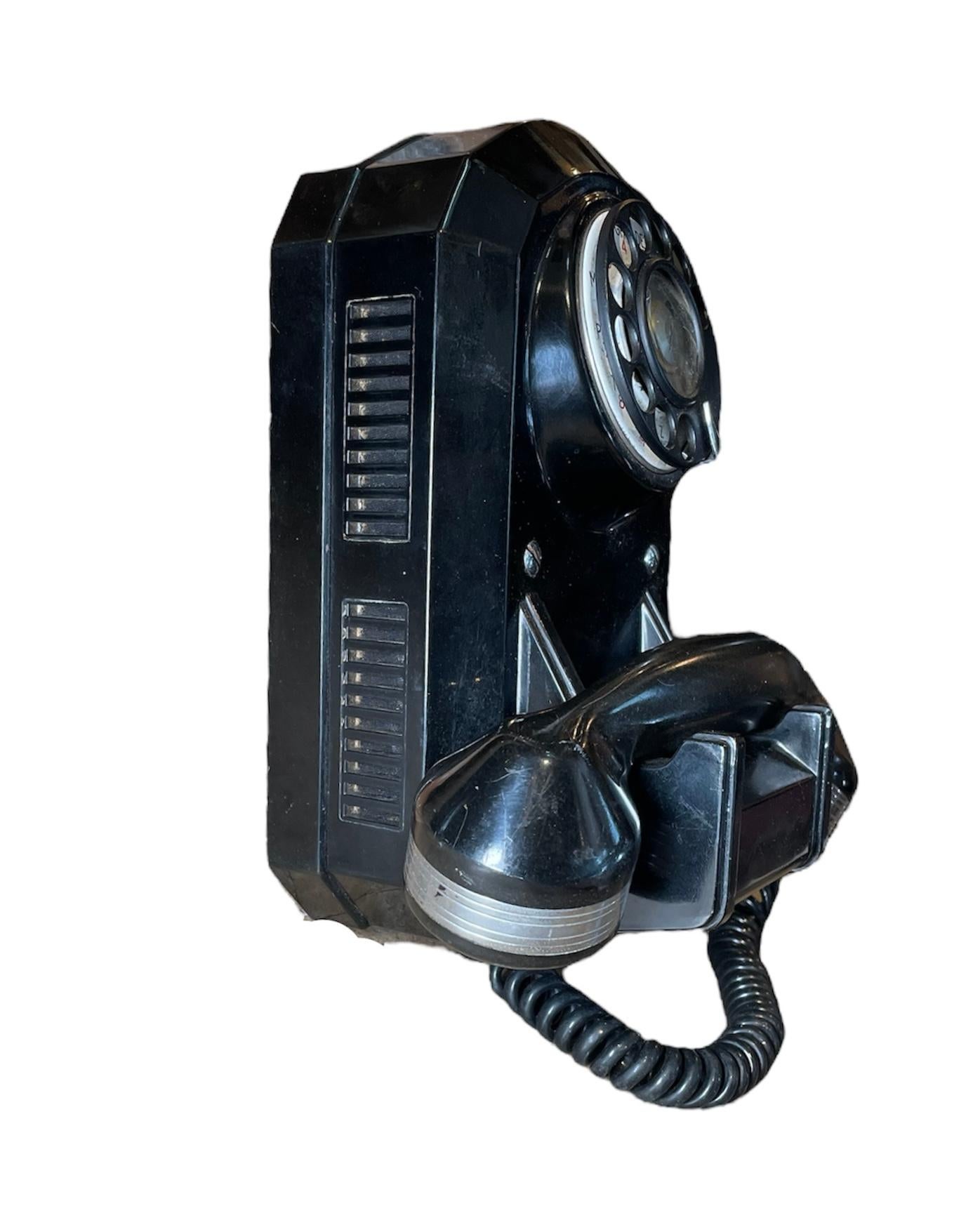 This is a Wall Mounted L 5000 Rotary Jukebox Telephone. It depicts a black color Bakelite tombstone shaped rotary telephone. In the back is hallmarked L 5000 BO, PE and ?N.S.4.